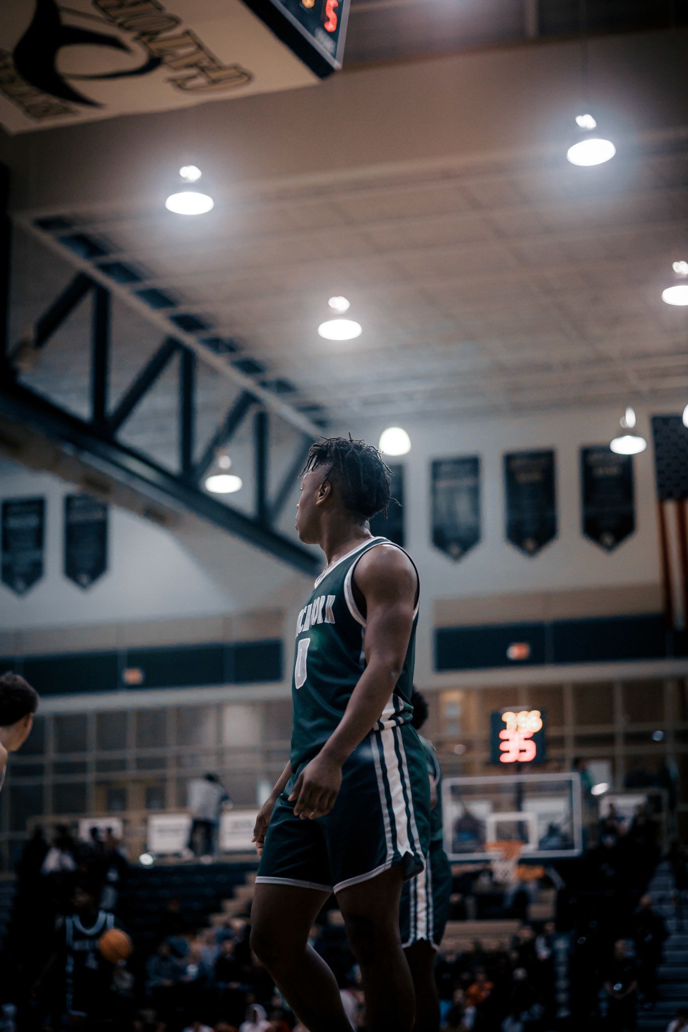 Basketball player in dark jersey stands on court with championship banners visible overhead.