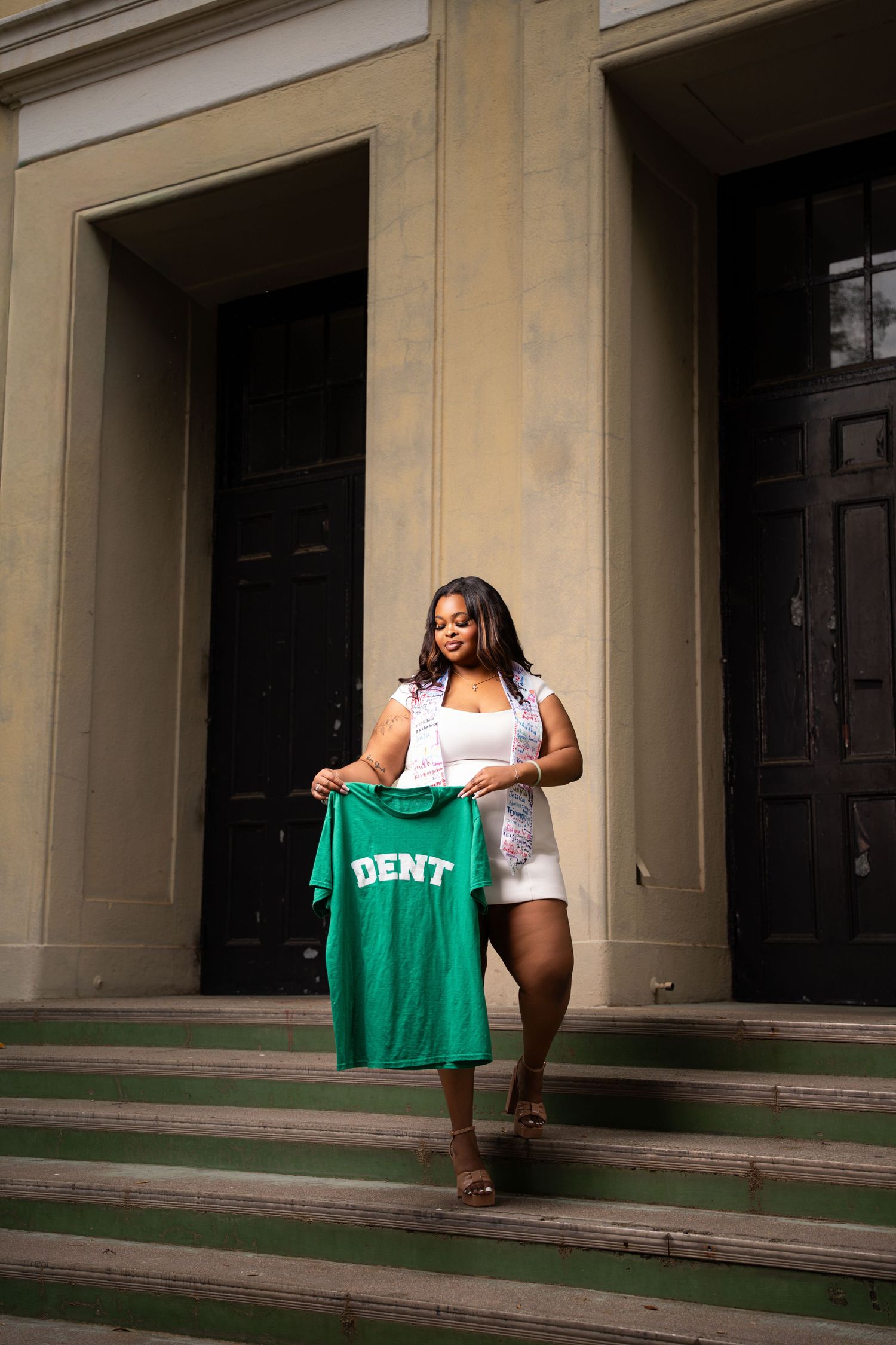 Young woman holding a green DENT t-shirt on building steps, wearing white dress and floral top.