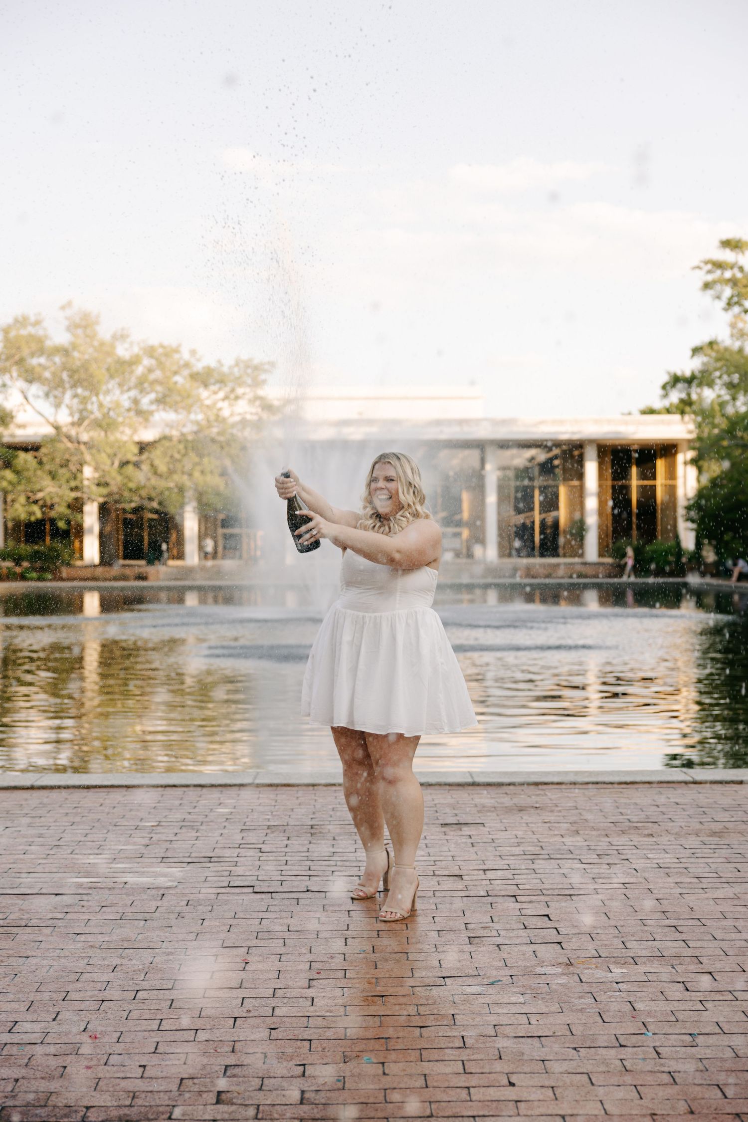 Woman in white dress popping champagne bottle by a fountain outdoors in golden hour light.