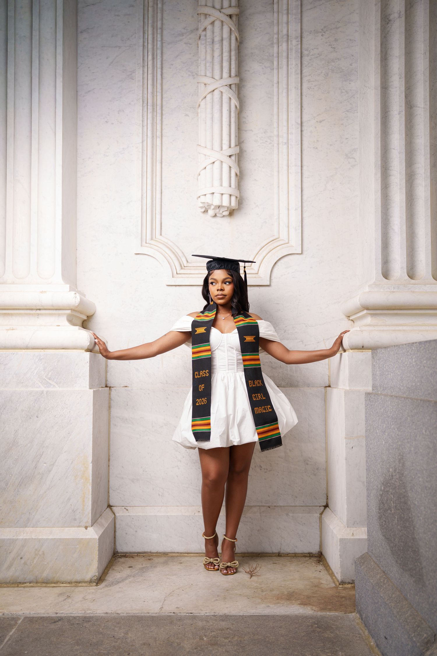 A young Black woman in a white dress, graduation cap, and kente cloth stole poses between marble columns.