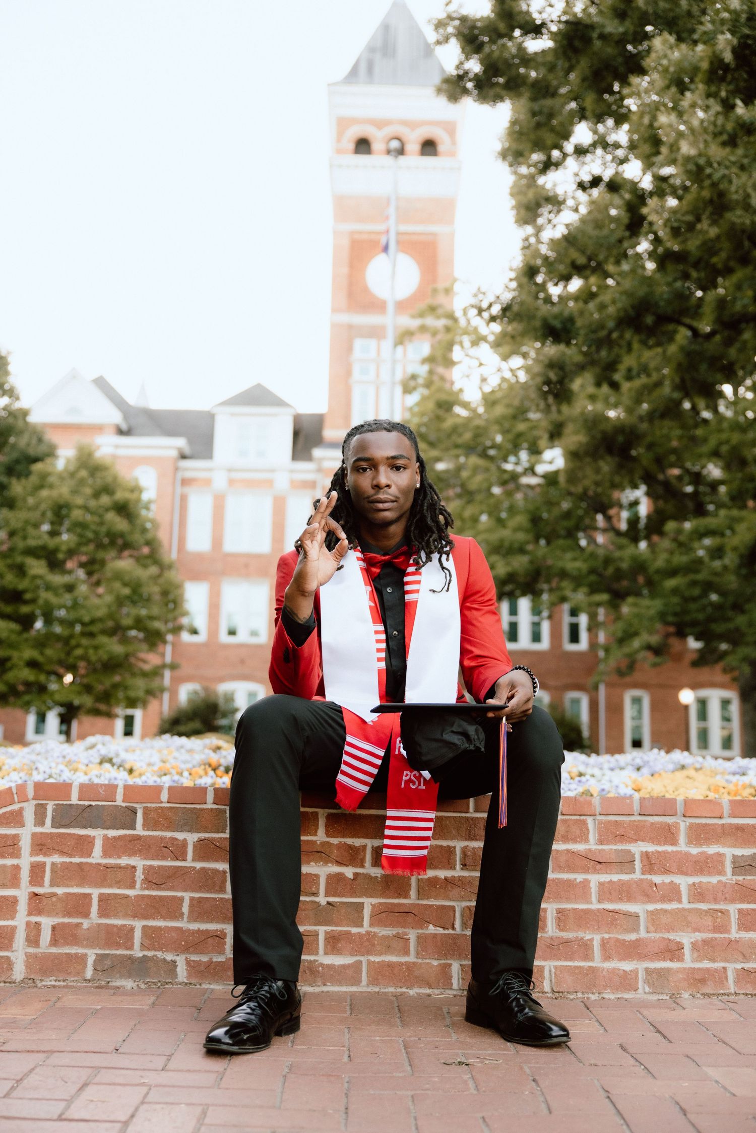 Graduate in red jacket and stole sits on brick wall in front of university clock tower building.