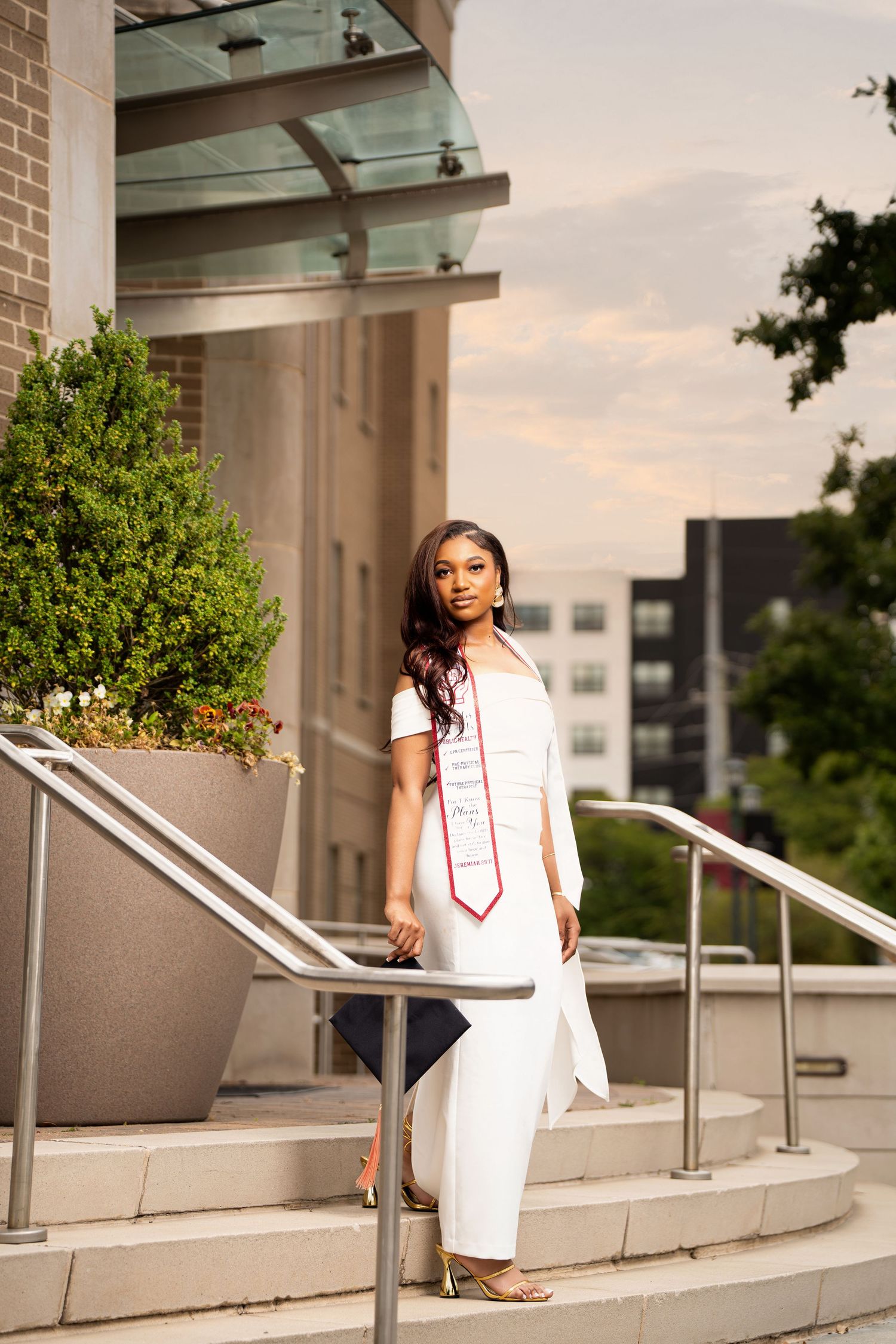 Young woman in white graduation dress and red stole poses on outdoor steps with urban backdrop at sunset.