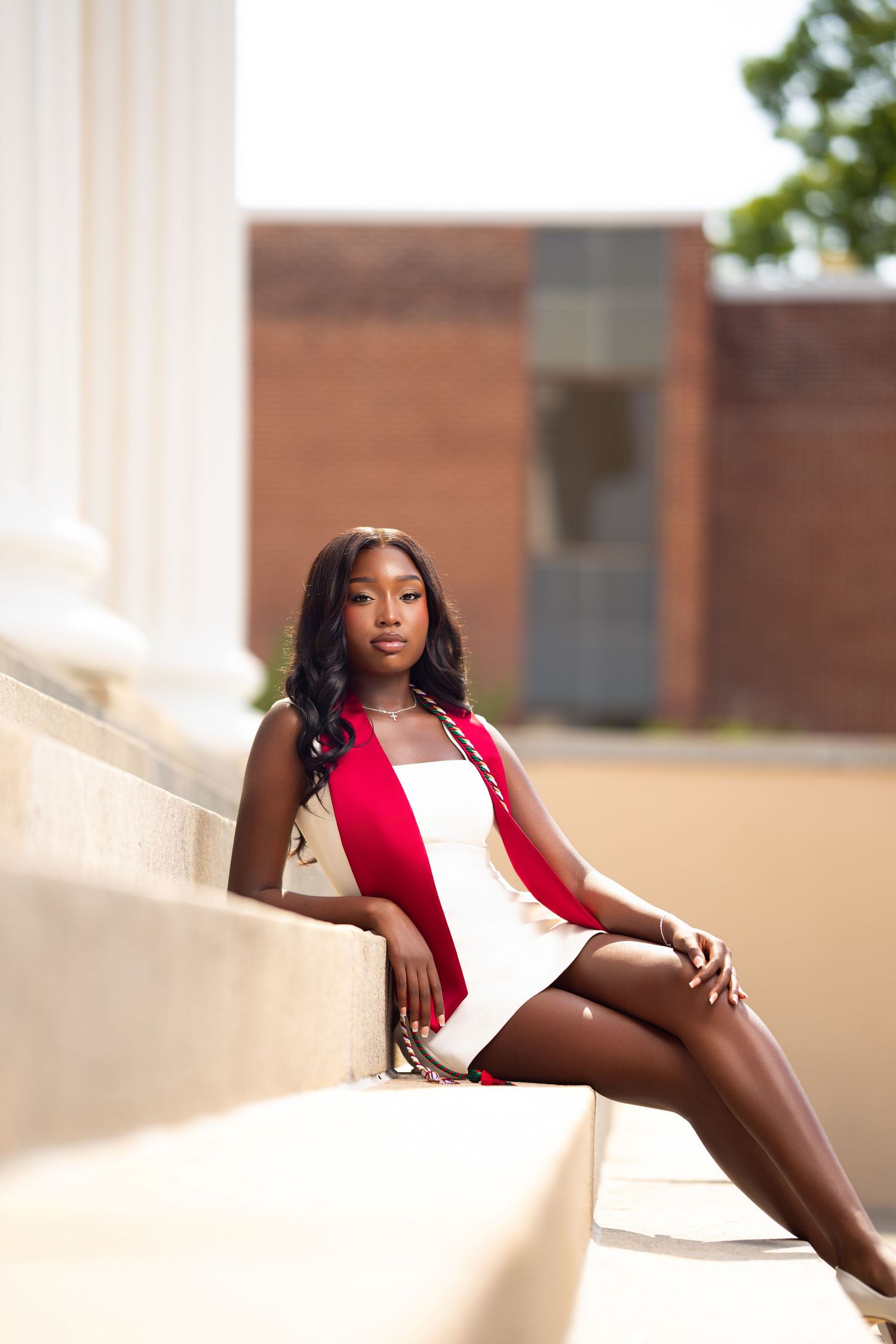 Young Black woman in red and white dress sitting gracefully on stone steps near a classical column outdoors.