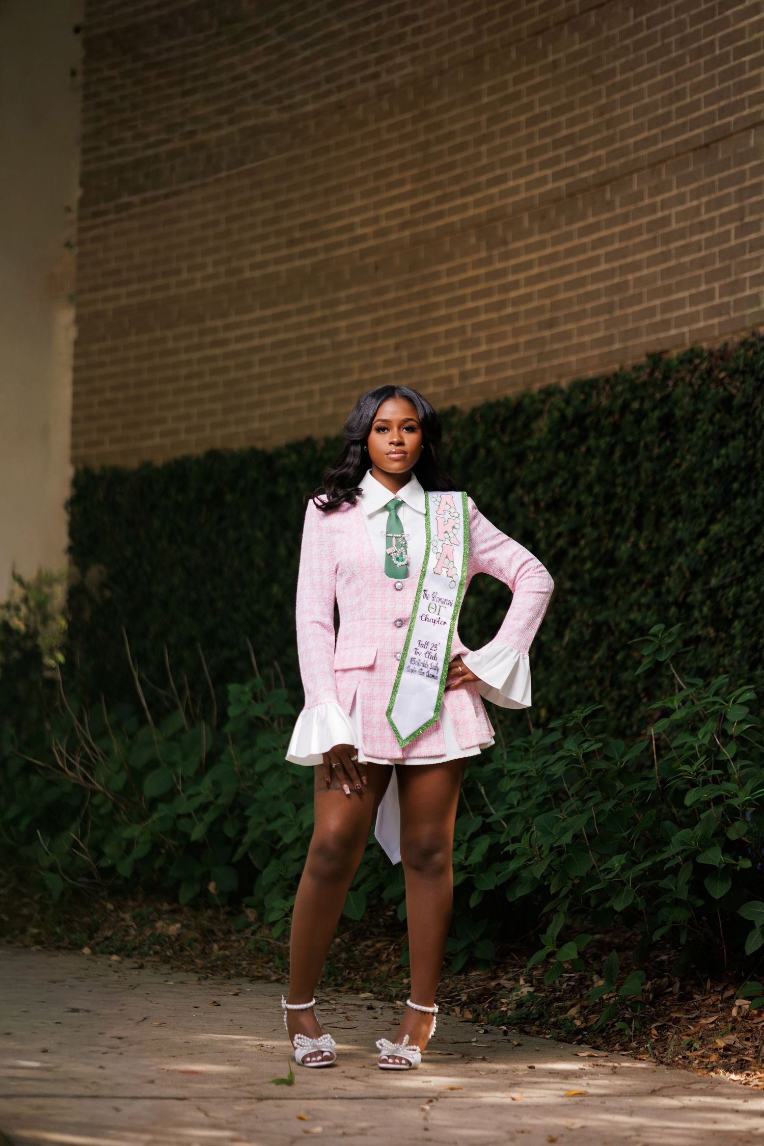 Young woman in pink plaid blazer dress with green sash poses confidently outdoors near brick wall and hedges.