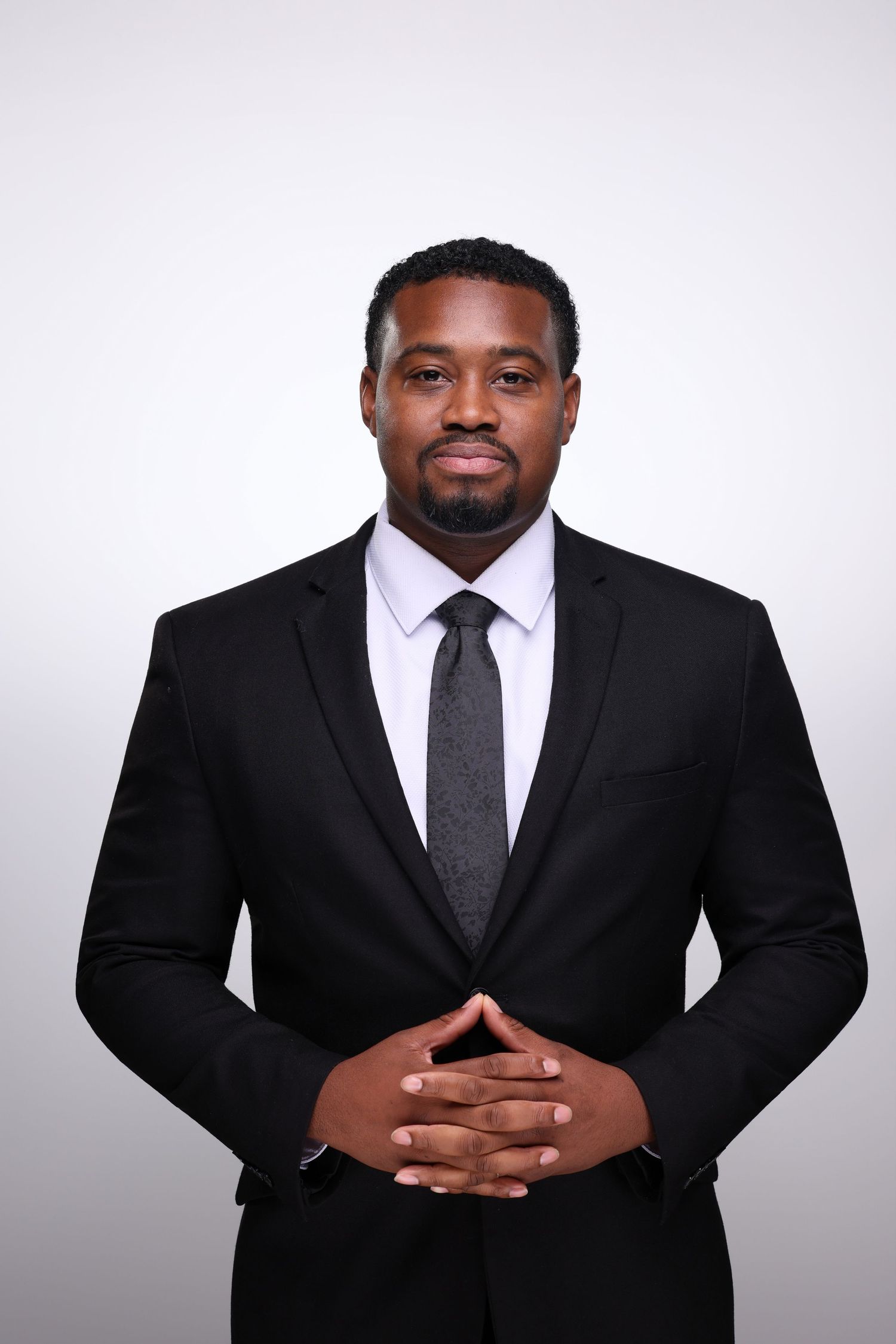Professional Black man in black suit and gray tie with clasped hands posing for a formal business portrait.