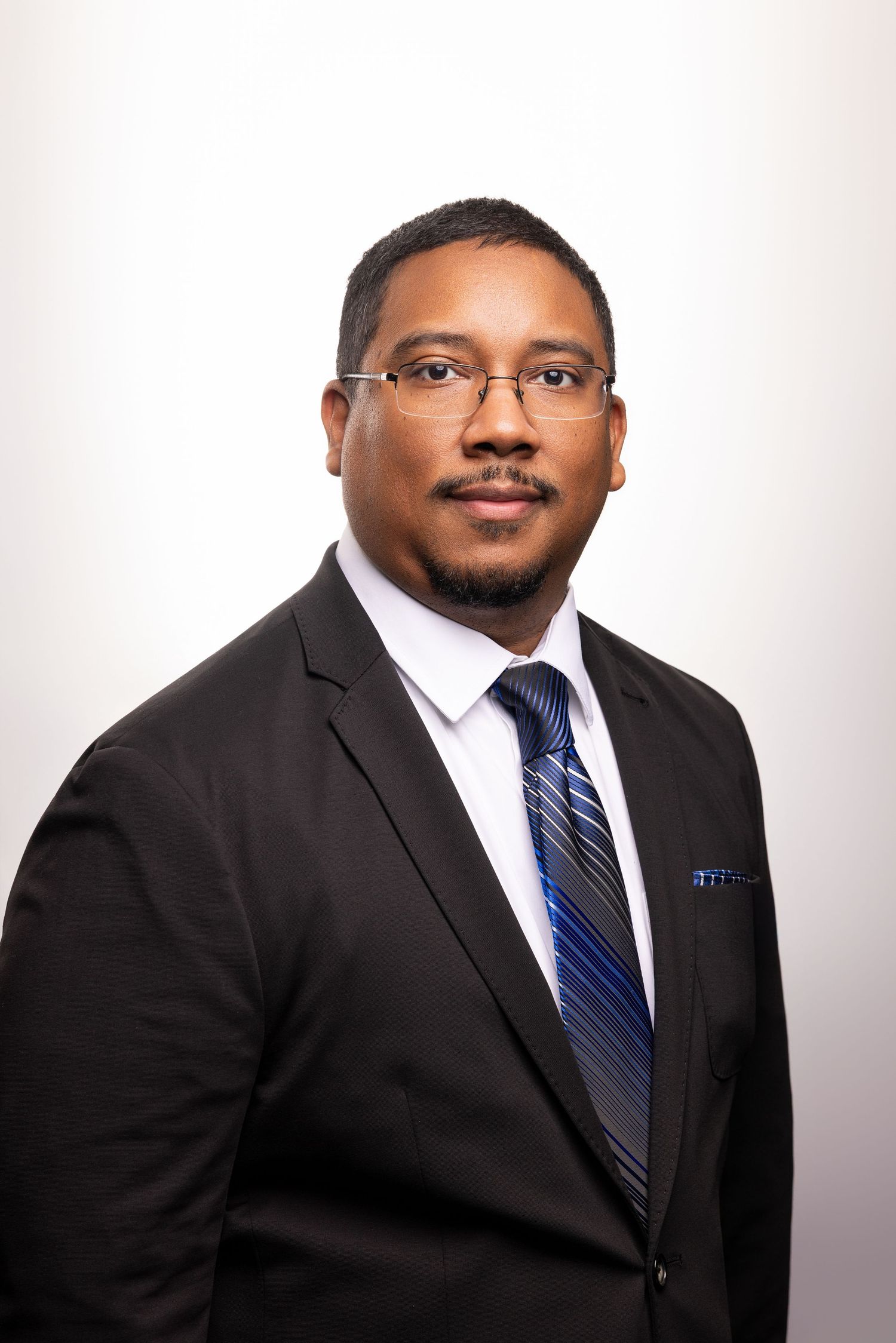Professional Black man in black suit, blue striped tie, and glasses poses for a formal business portrait photo.