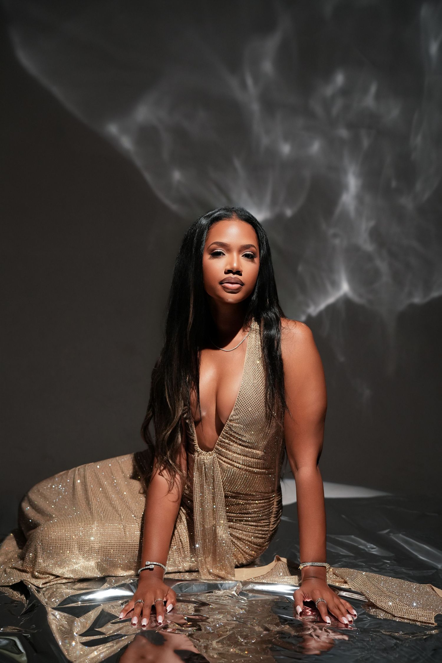 A woman in a gold glittery gown poses on a reflective surface with dramatic smoke effects in a dark studio.