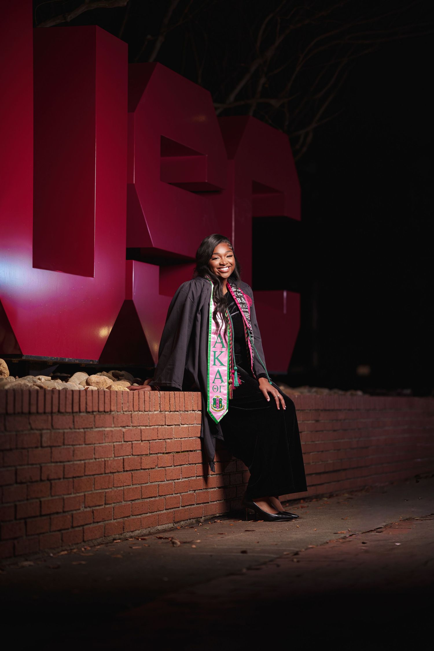 Graduate in black cap and gown with decorated stole stands against red wall during nighttime graduation ceremony.