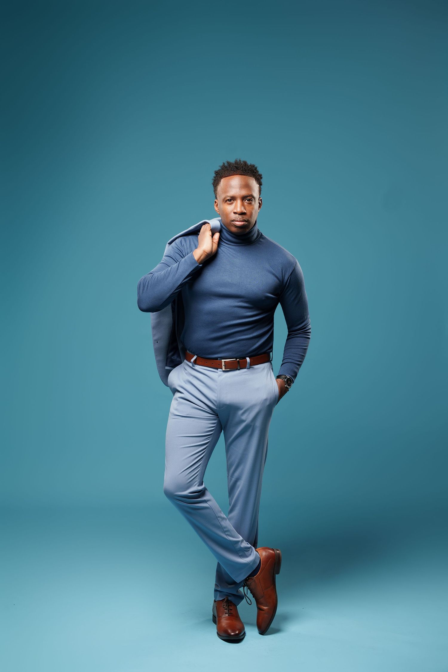 Professional fashion model poses in blue turtleneck and light pants against turquoise studio backdrop.