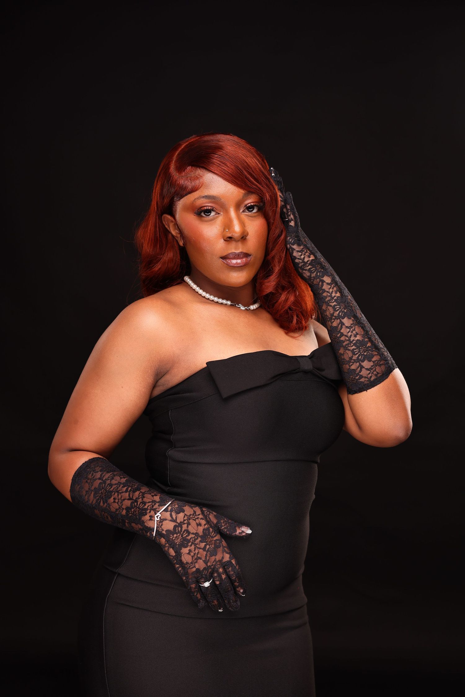 A figure in a black strapless dress and lace gloves poses dramatically against a dark background.