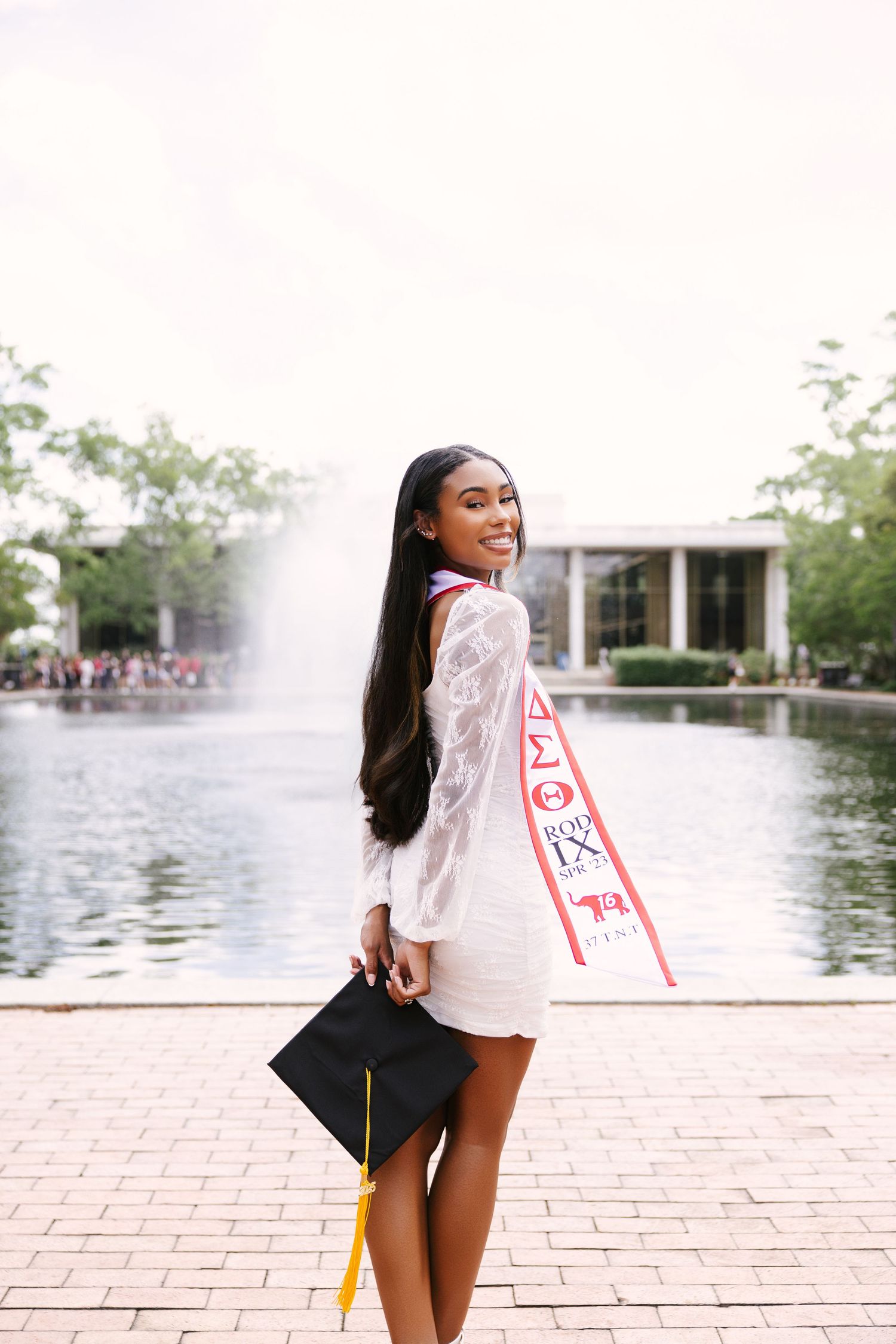 Graduate in white dress poses with diploma and graduation sash by fountain on campus during commencement celebration.