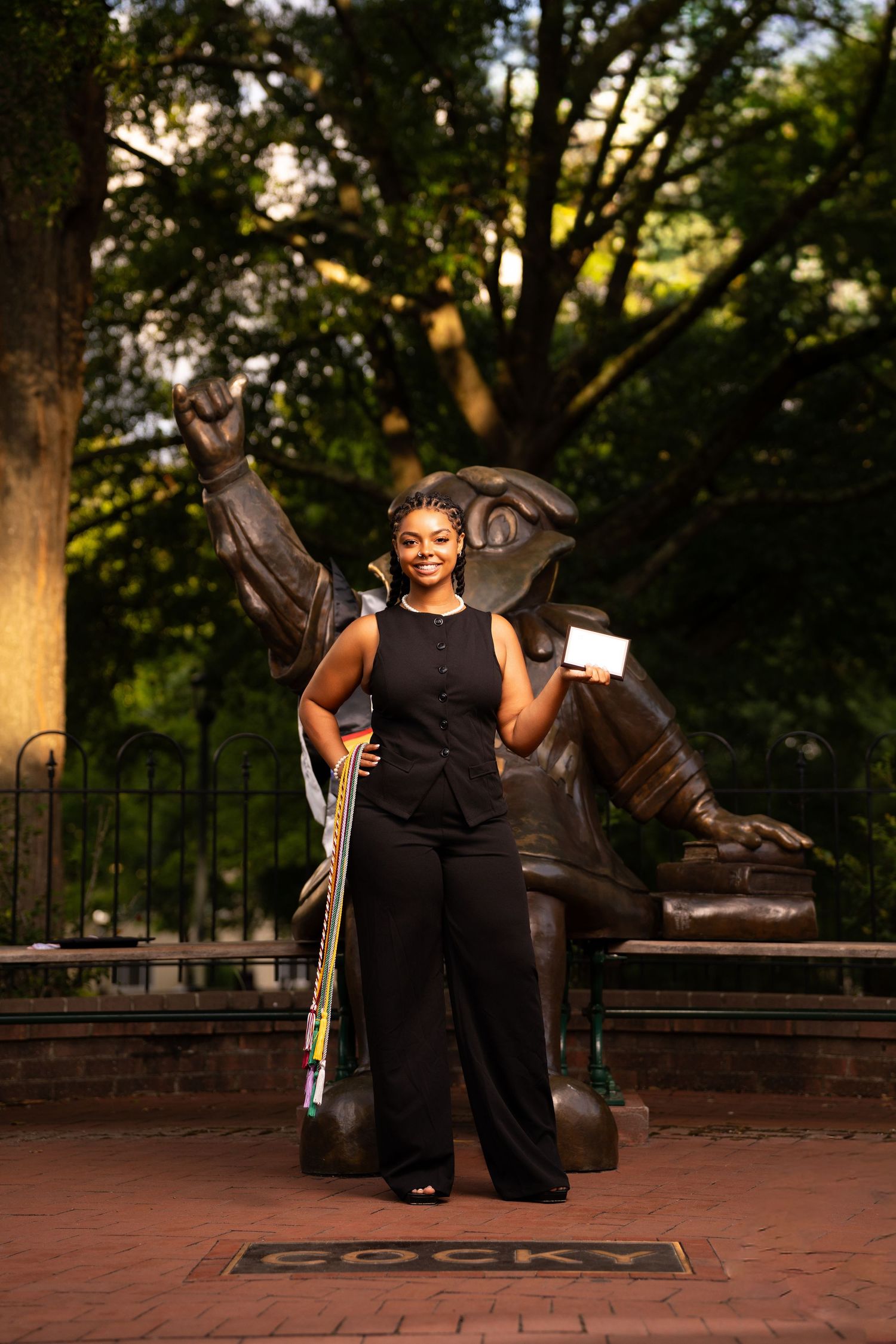 Graduate poses proudly in black outfit holding diploma beside Cocky mascot statue outdoors.