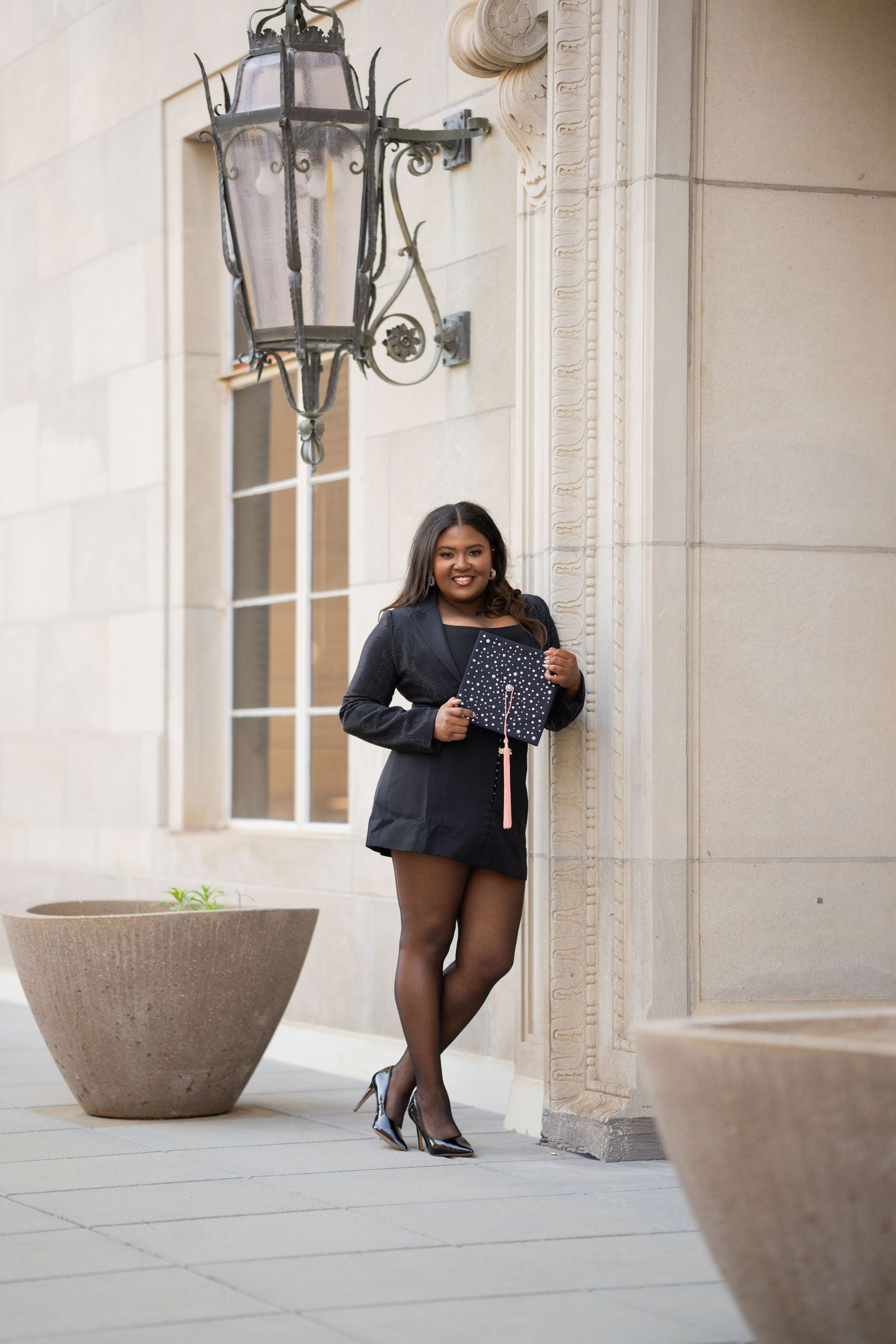 Young woman in black blazer holding graduation cap, leaning against stone building column with decorative lantern overhead.