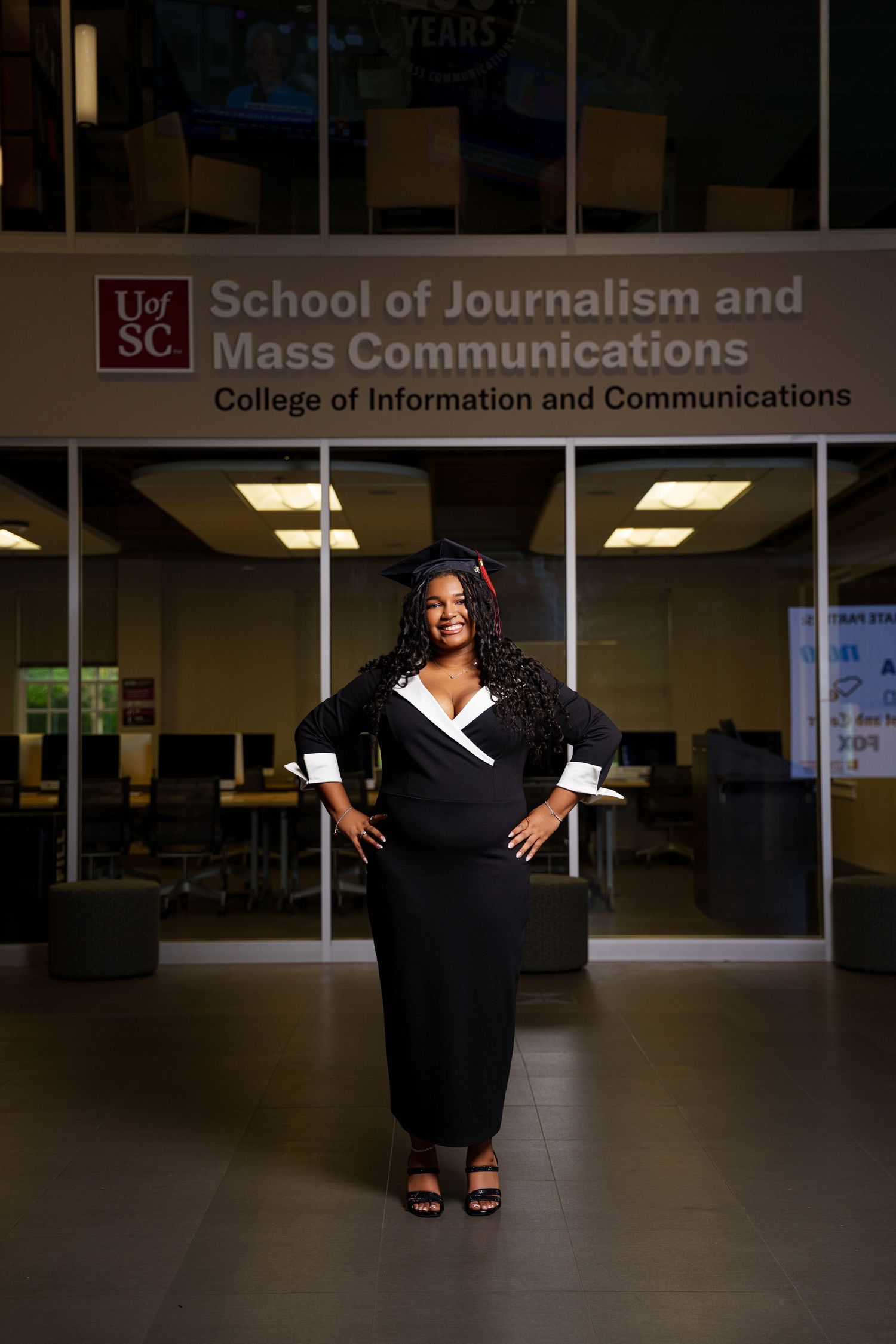 Graduate in cap and gown poses proudly outside UofSC School of Journalism and Mass Communications building.
