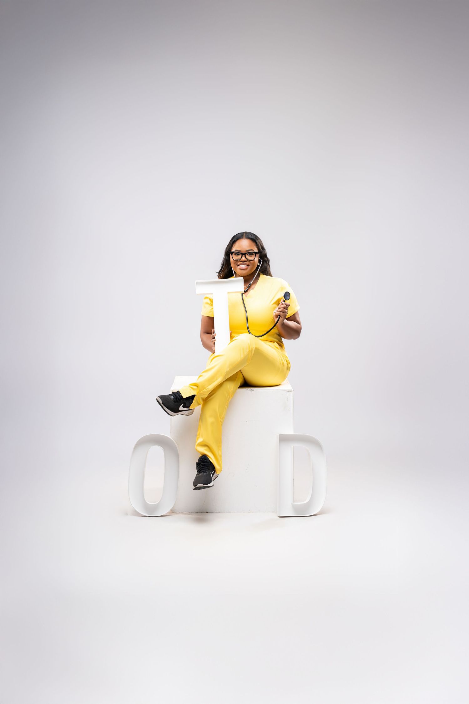 A woman in a yellow jumpsuit and glasses sits confidently on white block letters against a gray background.