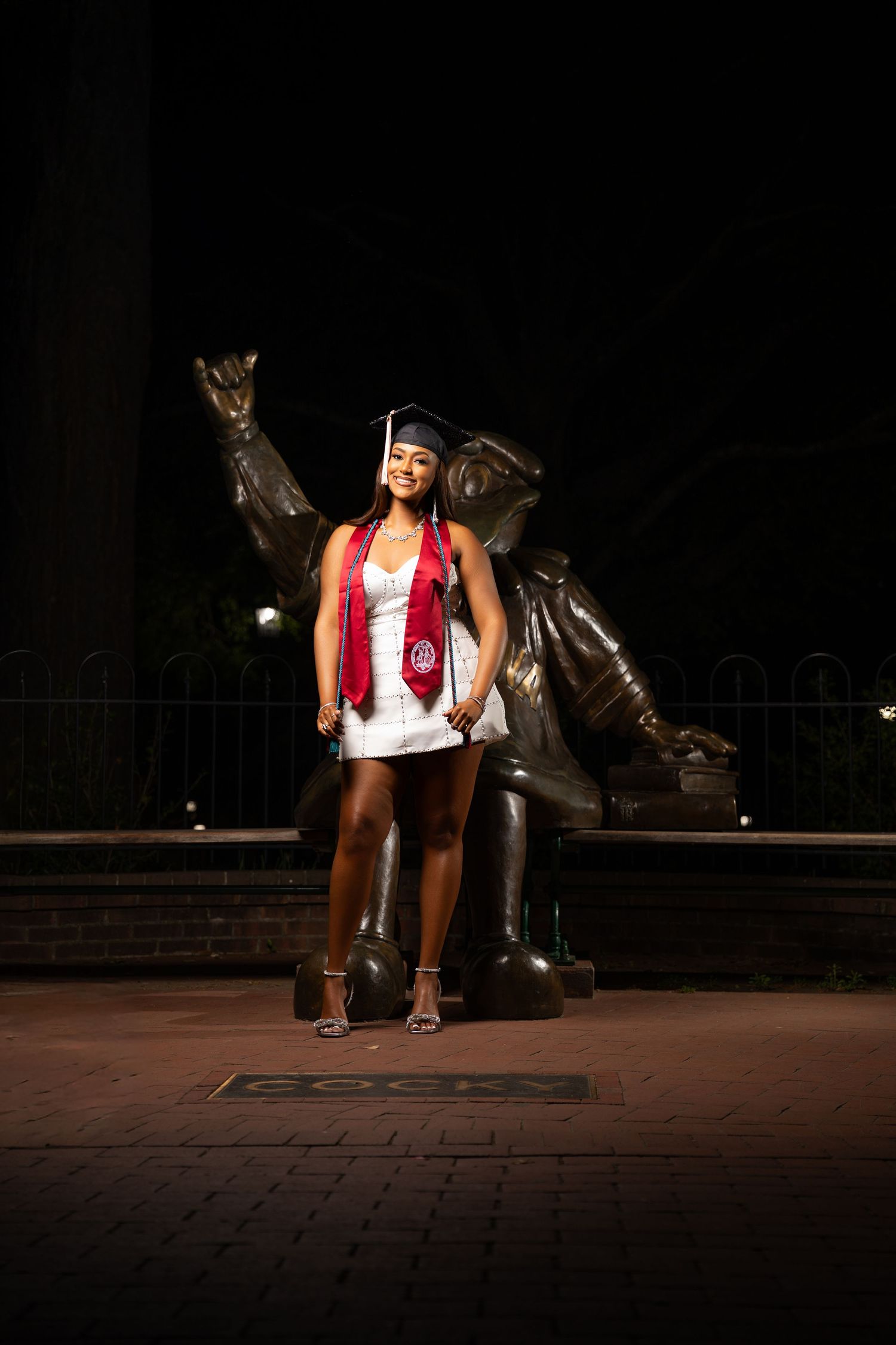 Graduate in cap and gown poses proudly in front of a bronze statue at night during commencement celebration.