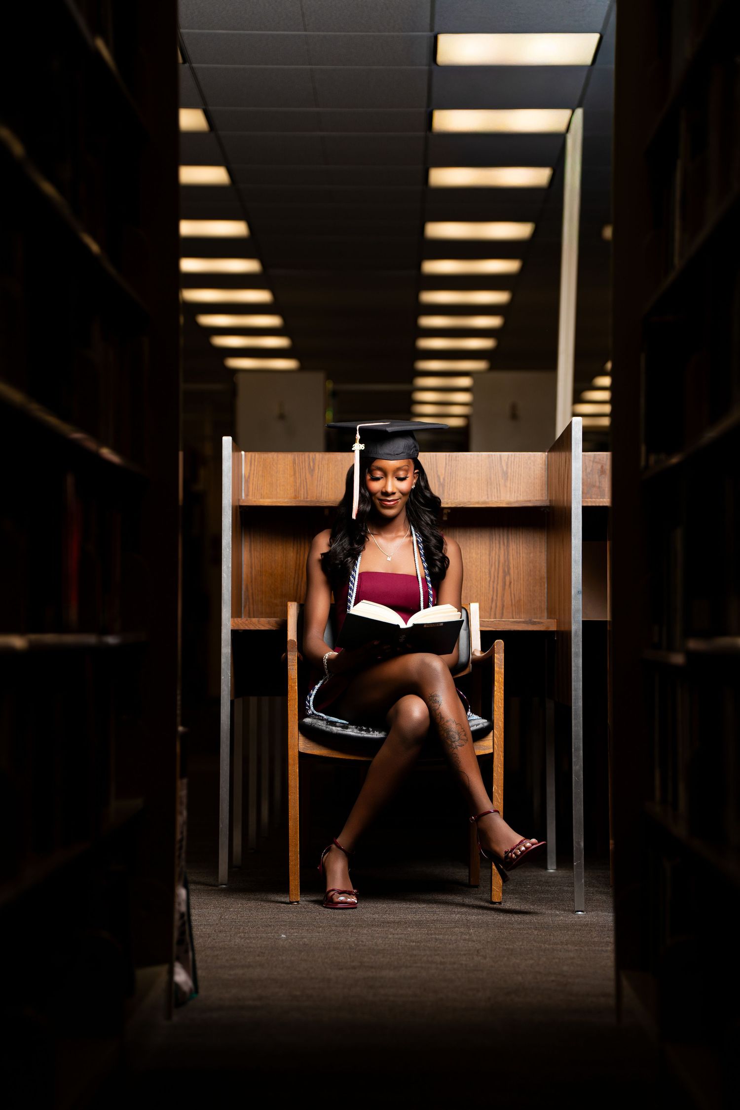 Young Black woman in graduation cap reads book at library study carrel, wearing maroon dress and heels.