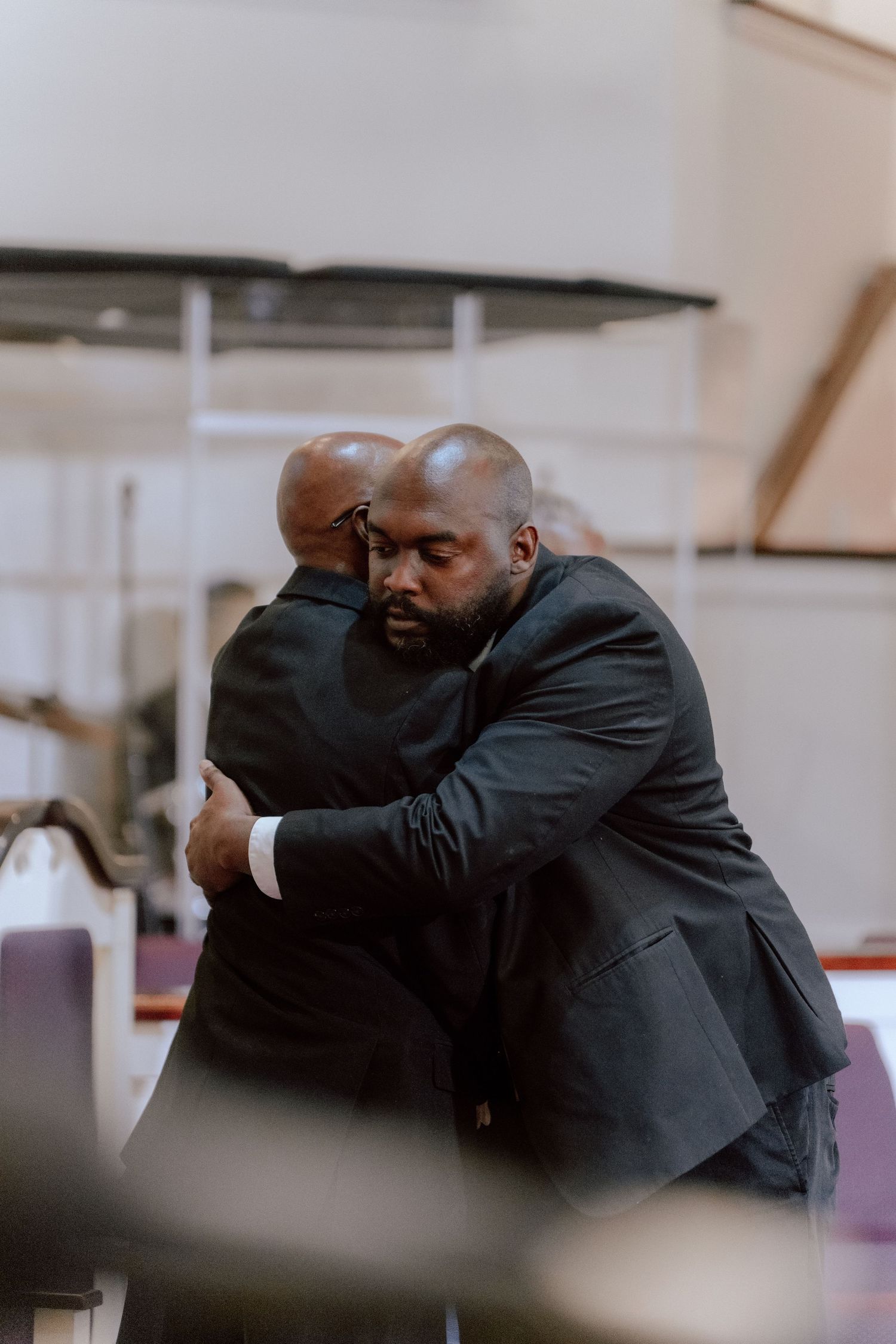 Two men in dark suits sharing a heartfelt embrace inside a church with purple pews.