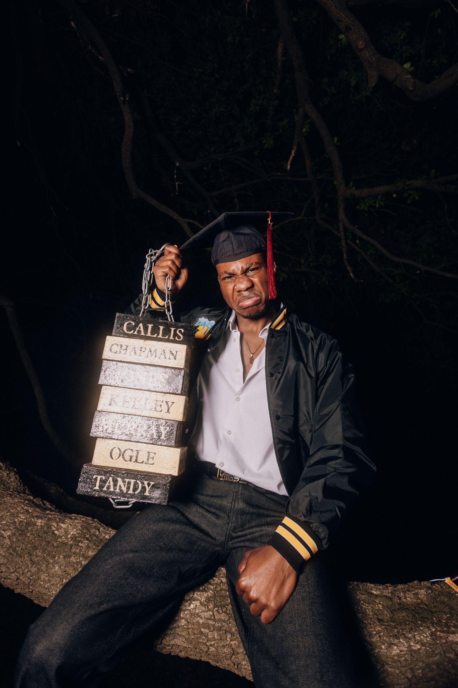 Graduate in cap and gown sits with stacked books in nighttime outdoor setting.