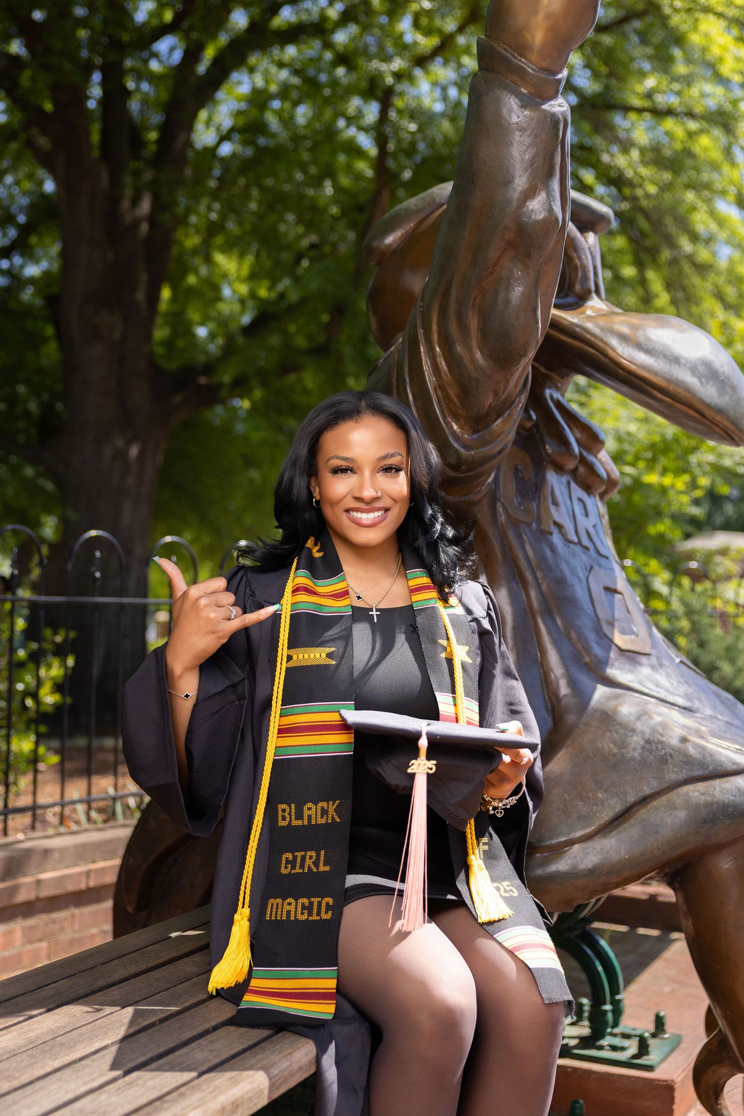 Graduate poses with graduation stoles and regalia near outdoor statue on college campus.