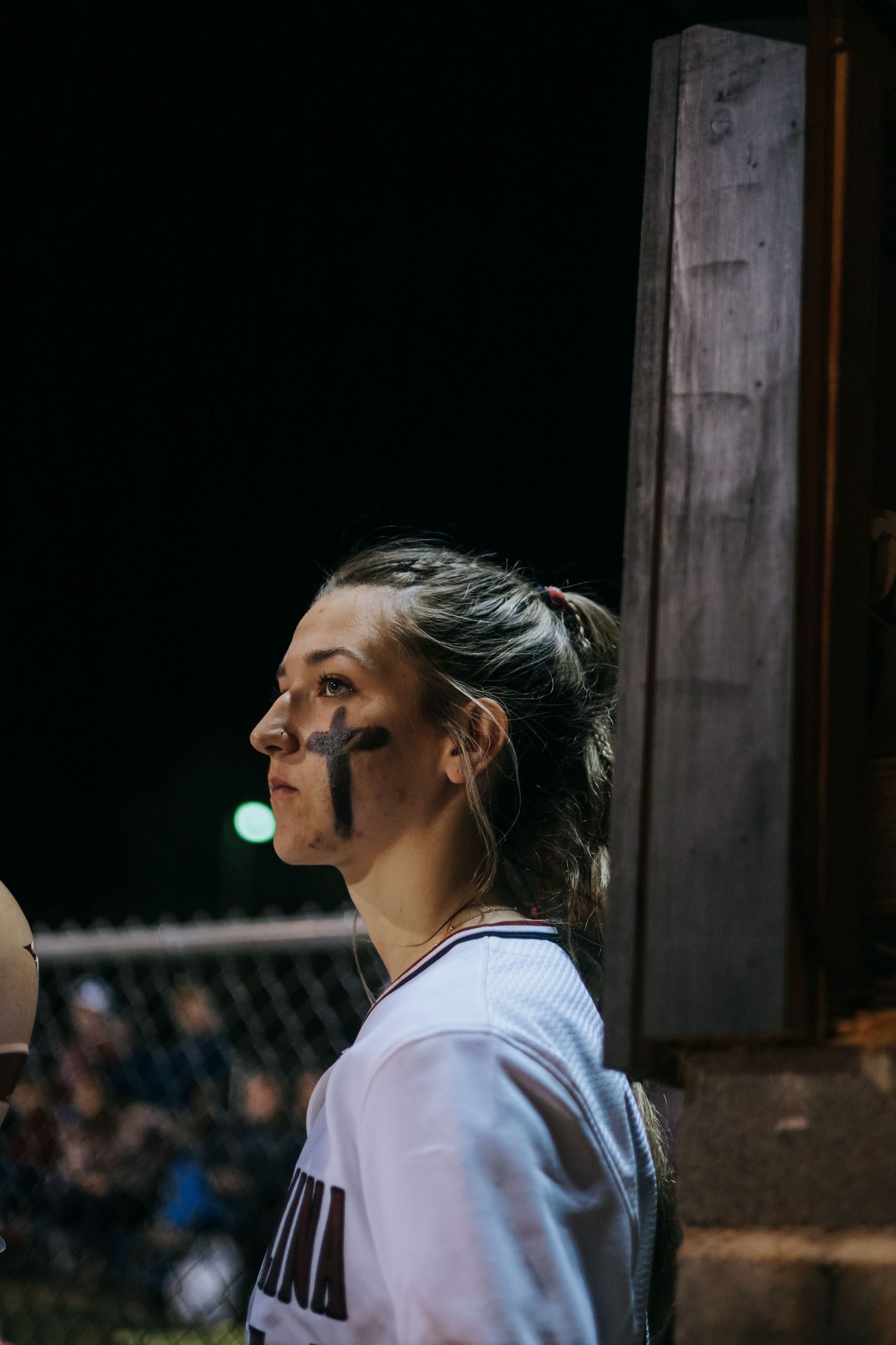 Softball player with eye black wearing white uniform in dugout at night.