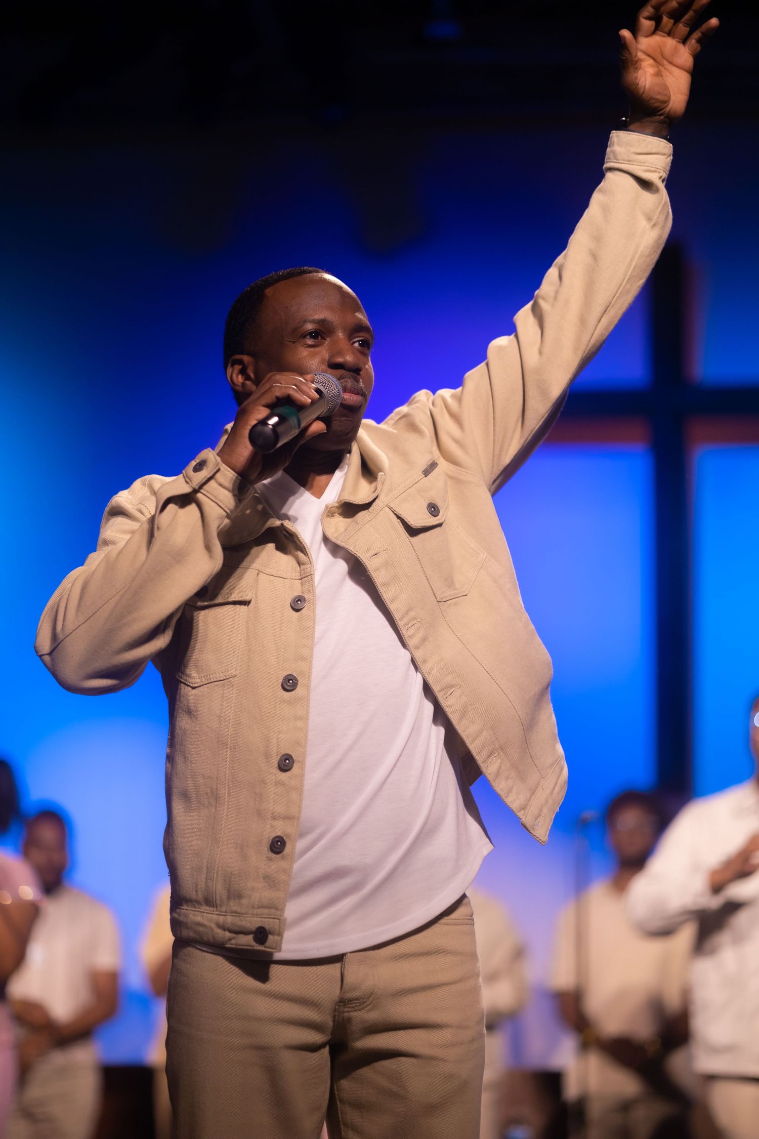 A performer in a tan jacket sings passionately with one arm raised during a gospel concert performance.