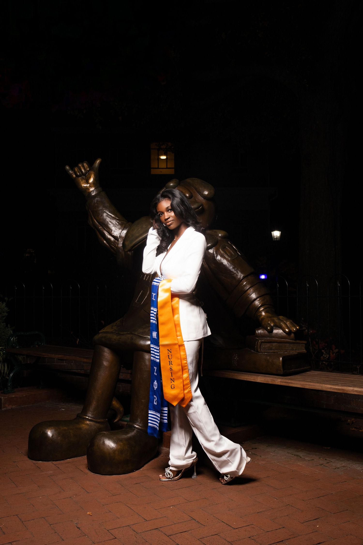 Person in white outfit posing with Disney statue at night while wearing graduation stole.