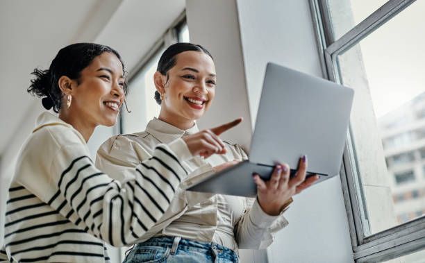 Two coworkers share a laugh while collaborating on a laptop by a bright office window.