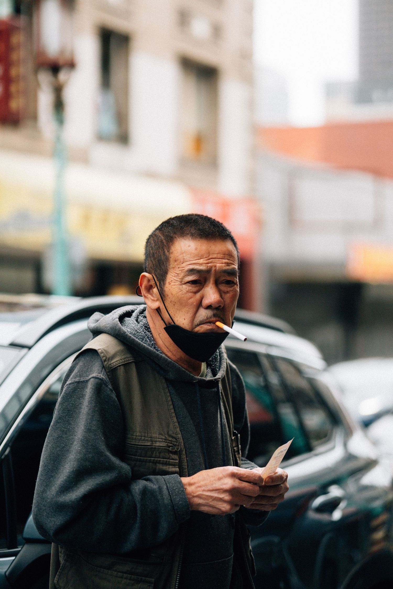 A person wearing a dark jacket looks at their mobile phone while standing near parked cars on a city street.