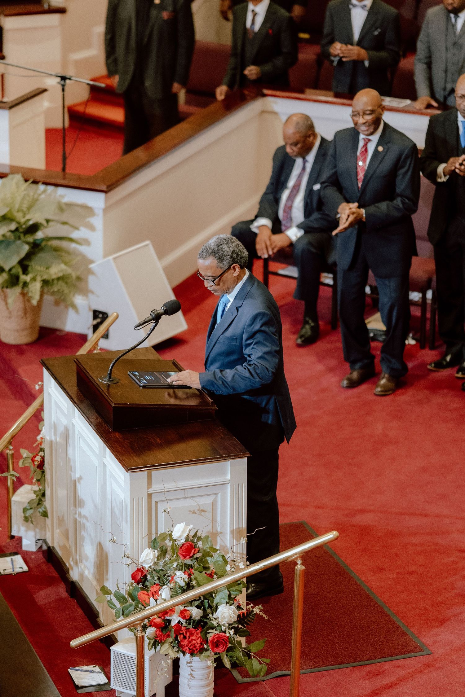 A speaker stands at a wooden pulpit with red carpet and floral arrangements in a church setting.