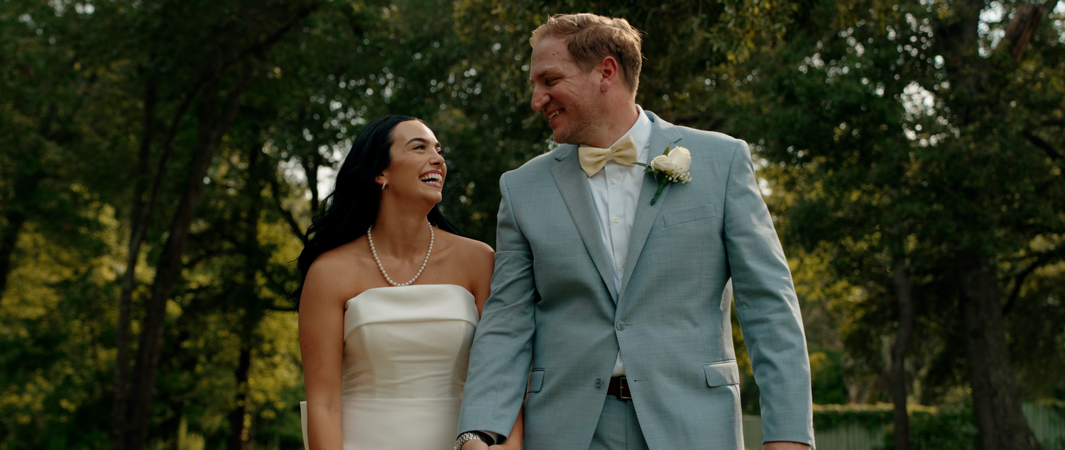 A couple in wedding attire walks together and smiles while surrounded by lush green trees in an outdoor setting.