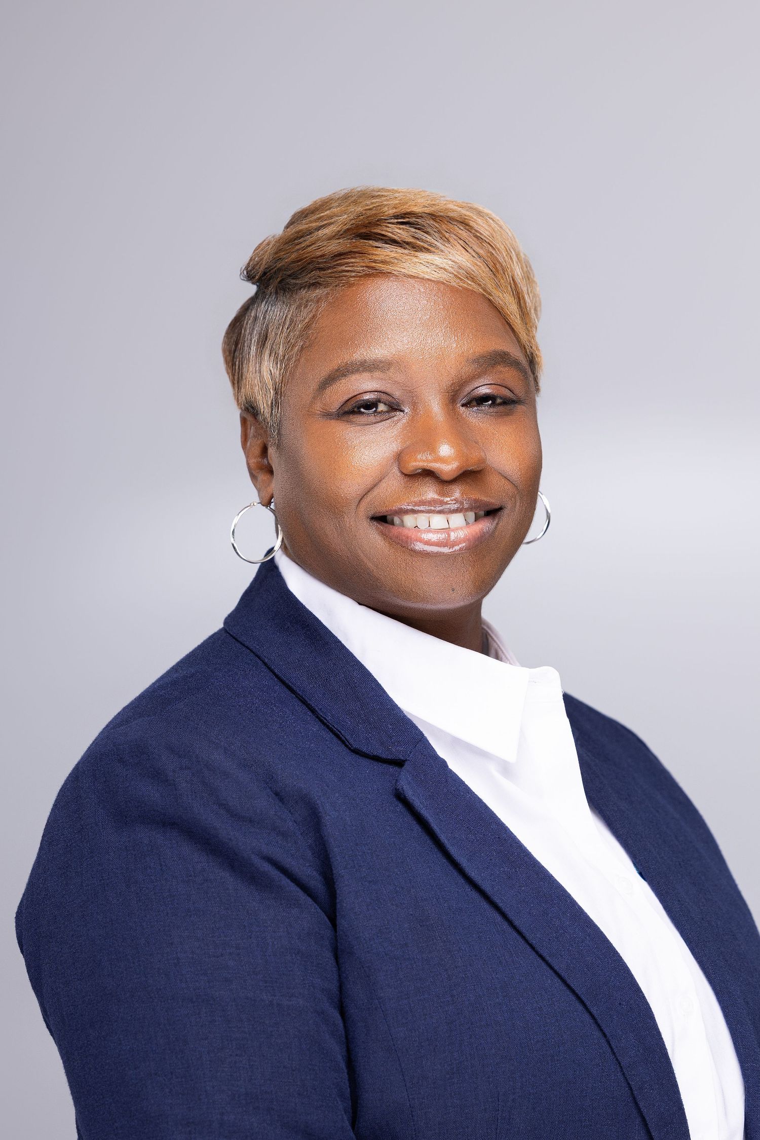 Professional headshot of a person wearing a navy blazer and white blouse, smiling warmly against a light gray background.