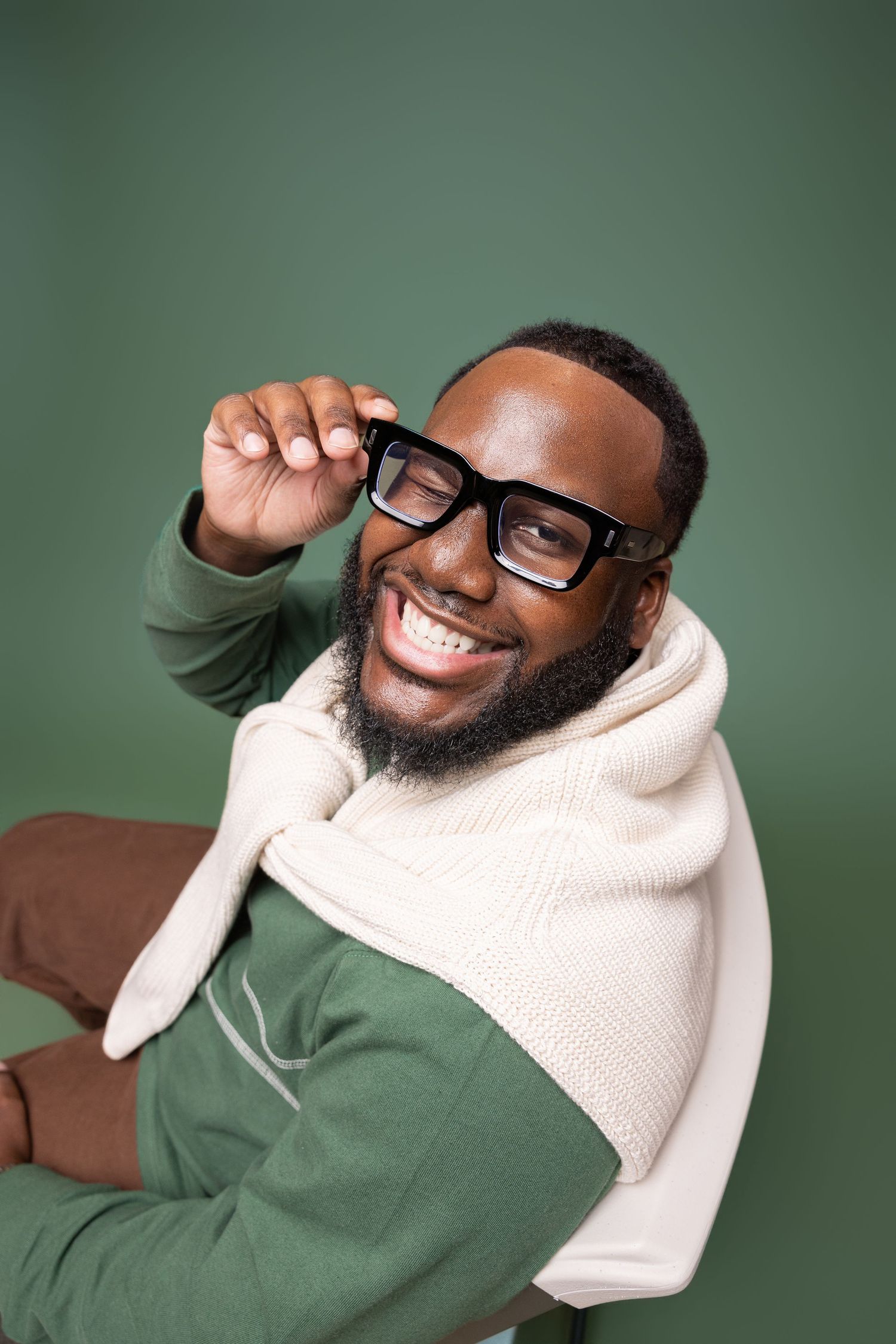 Smiling person wearing glasses, a cream-colored scarf and olive green sweater against a green background.