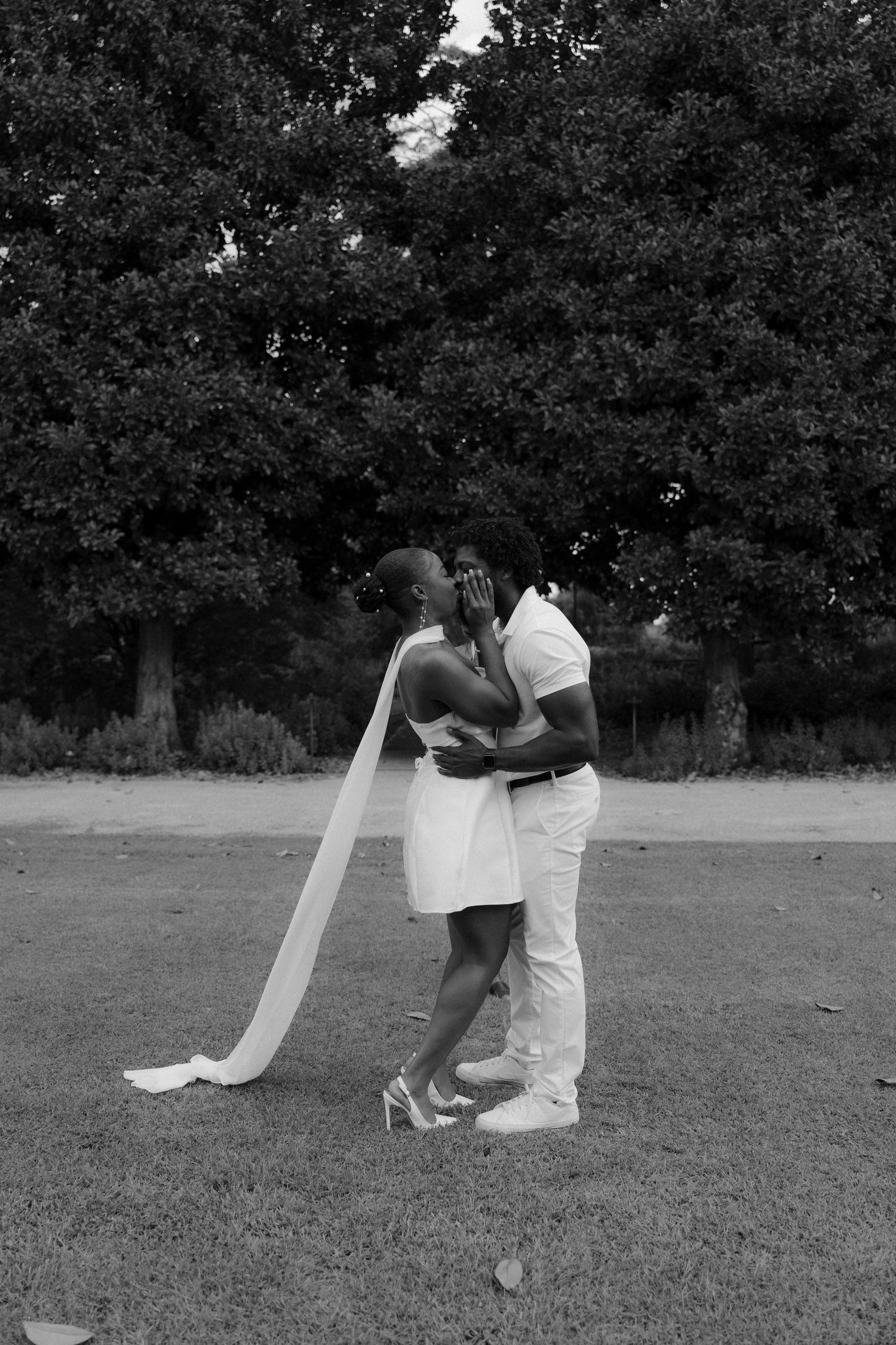 Black and white photo of a couple in white attire embracing on a lawn with lush green trees in the background.