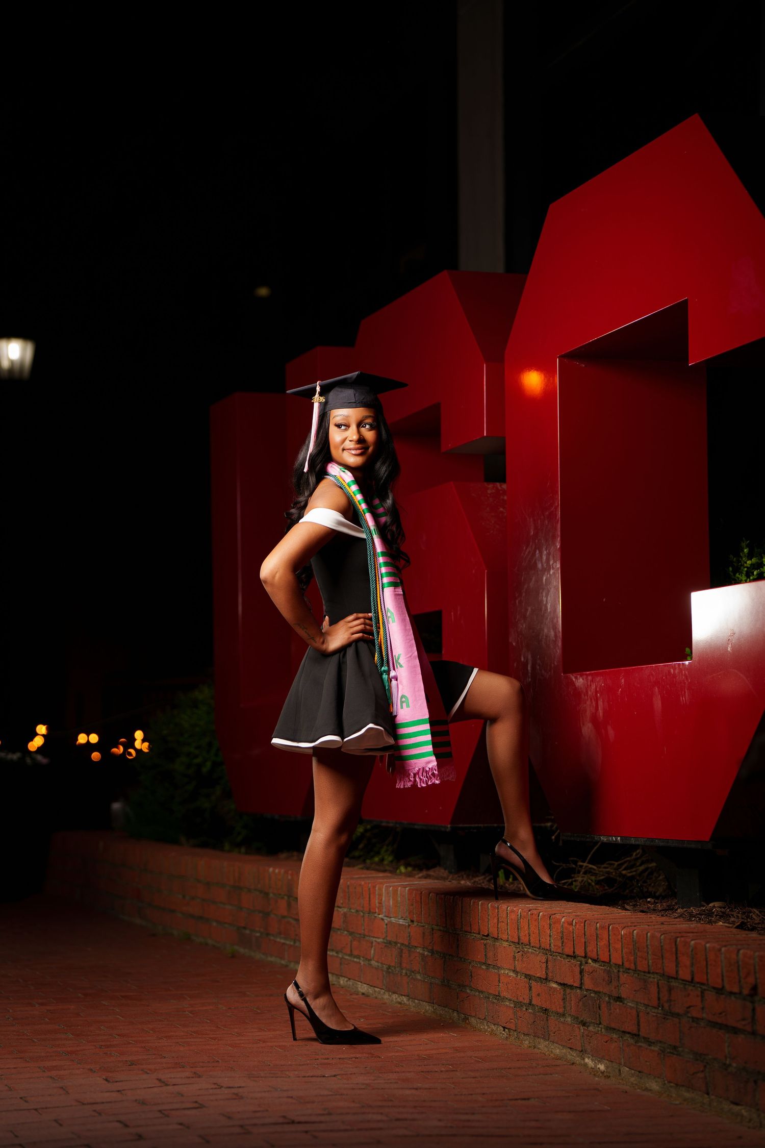 Young woman in graduation cap, gown, and colorful sash poses proudly at night near red university sign.
