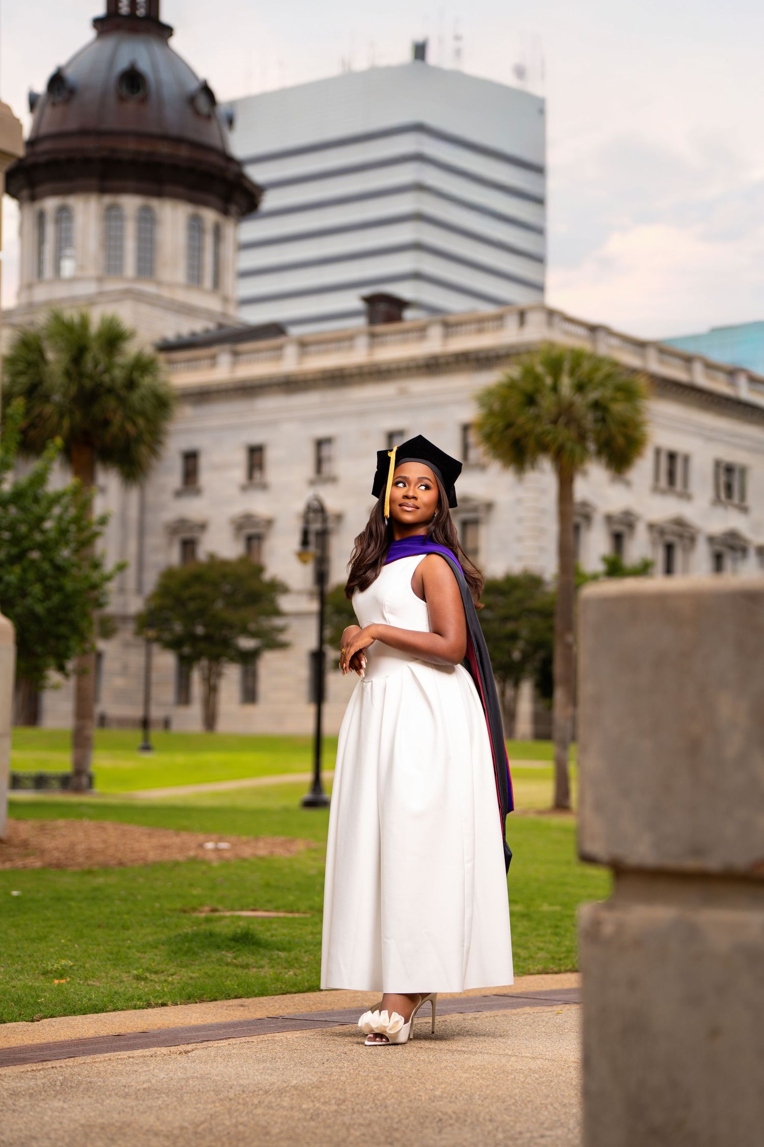 Young Black woman in white dress and graduation cap and gown poses outdoors near historic building with palm trees.