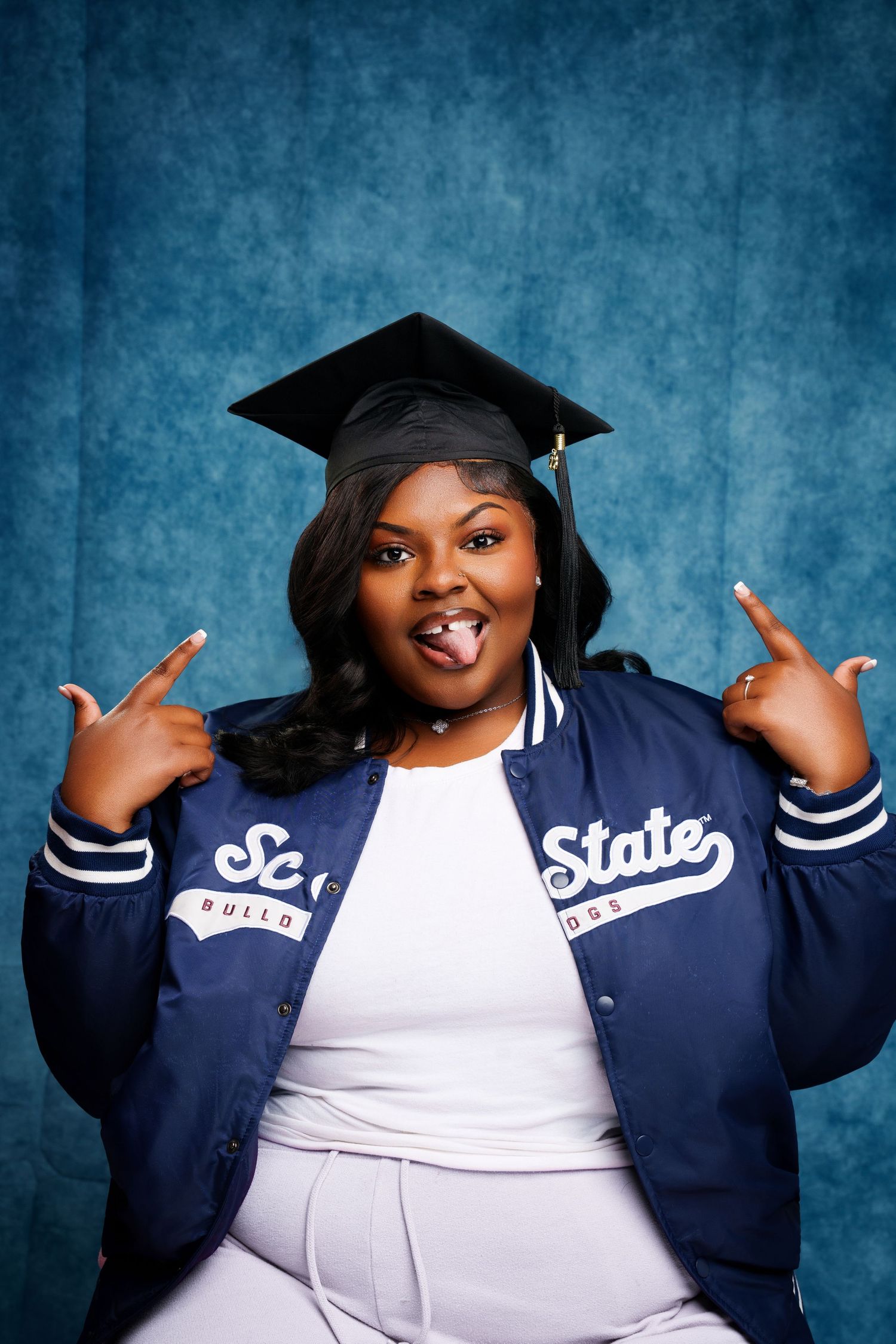 Excited graduate wearing cap and State varsity jacket, pointing fingers and sticking tongue out against blue backdrop.