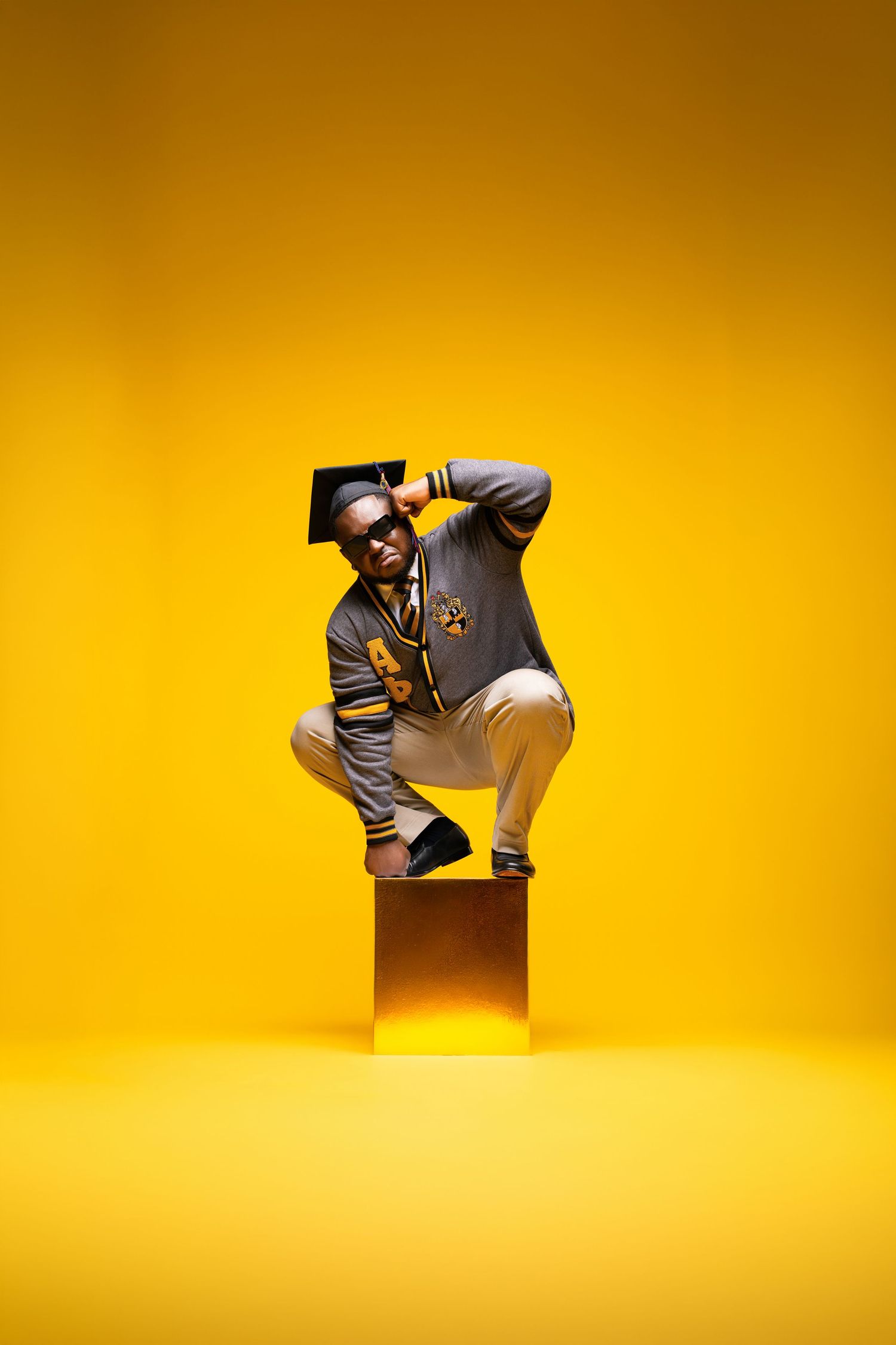 Man in graduation cap and varsity jacket squatting on gold cube against vibrant yellow background.