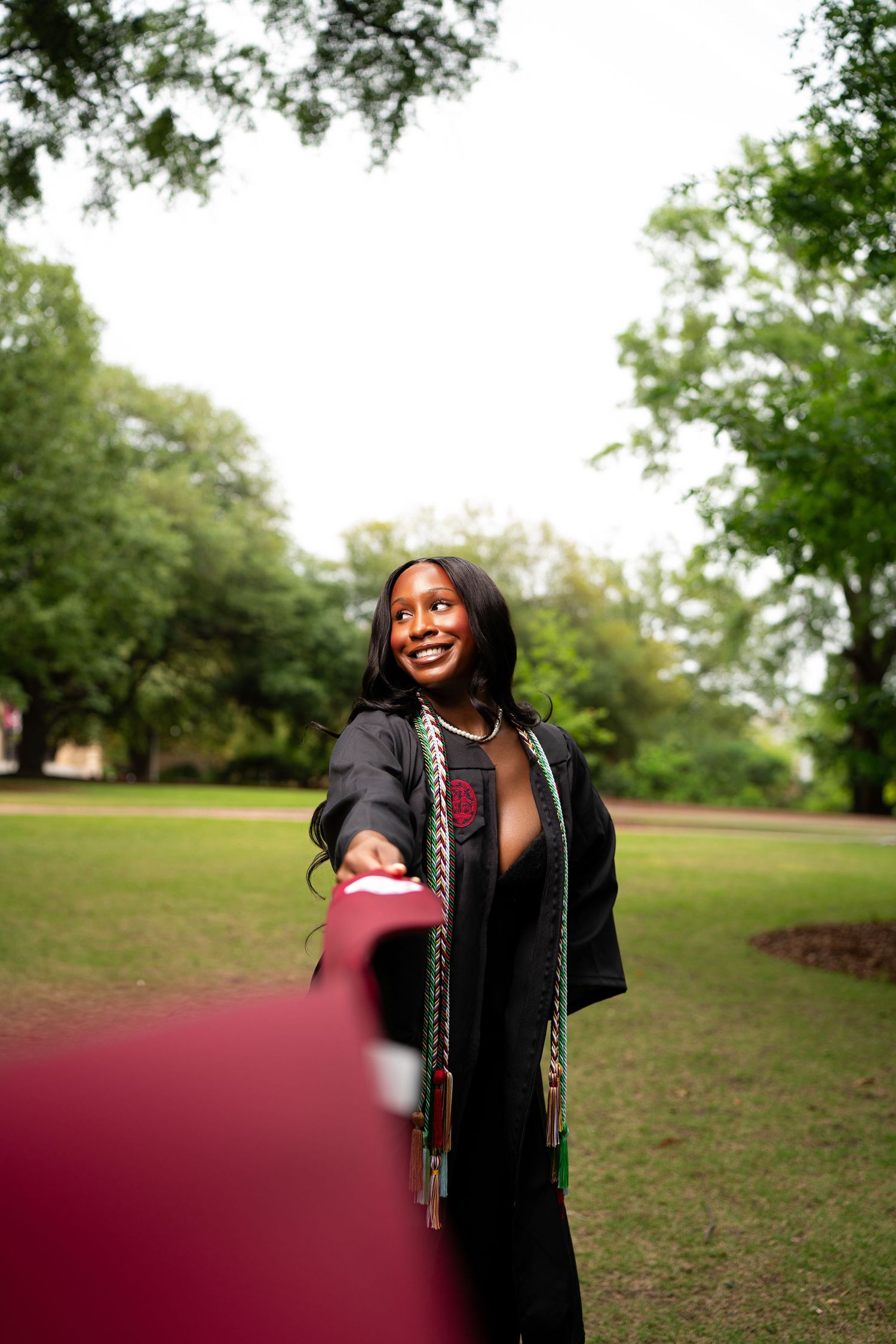 Smiling graduate in black gown with colorful cords reaches toward camera outdoors on green campus lawn.