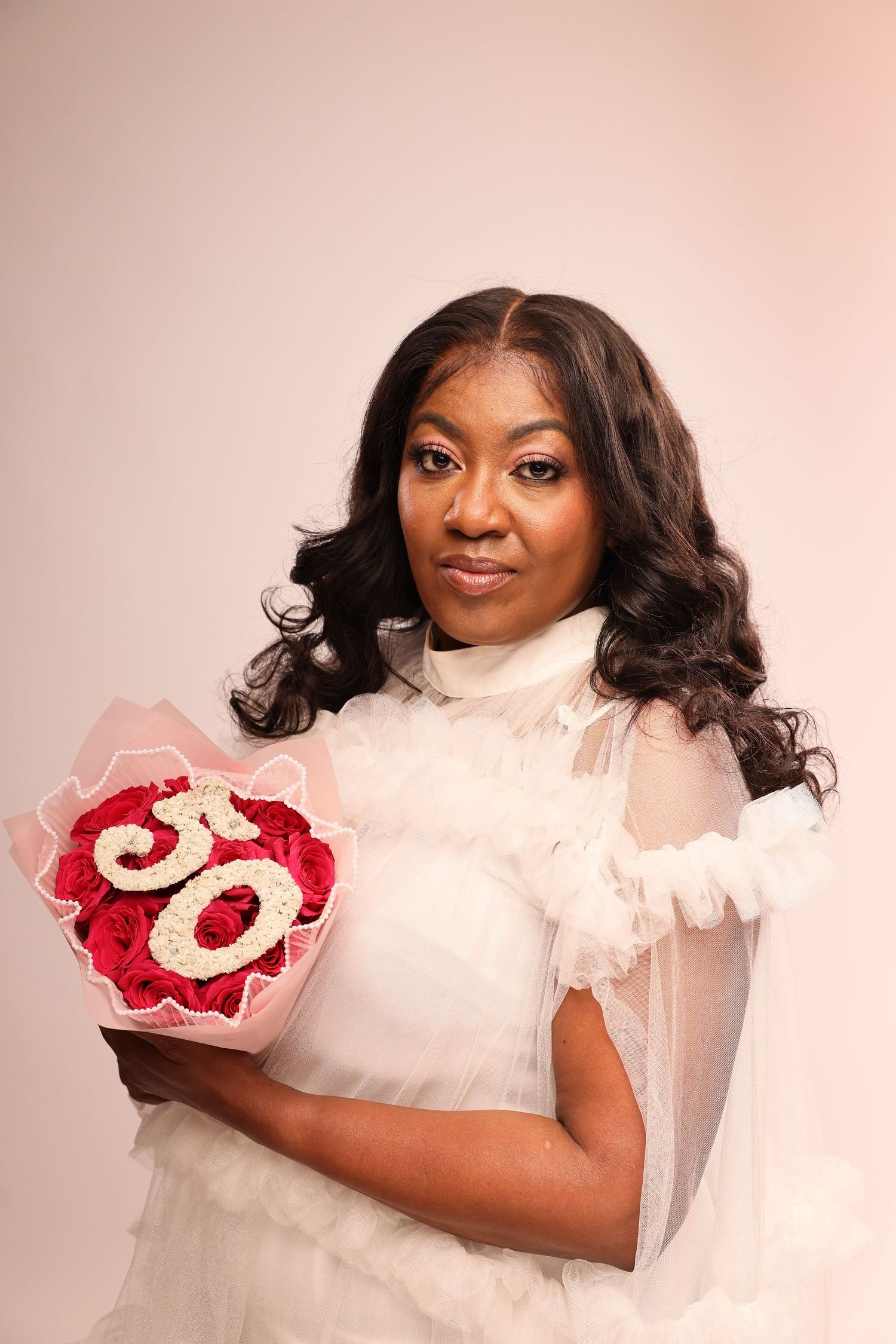 Portrait against pink backdrop showing person in white dress holding red and gold number 50 celebration decoration.