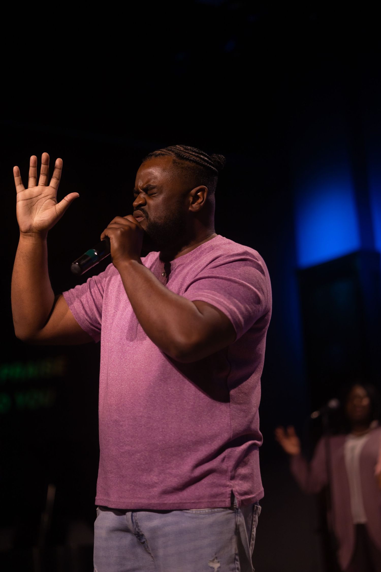 A performer in a pink shirt sings with raised hand against a blue-lit backdrop.