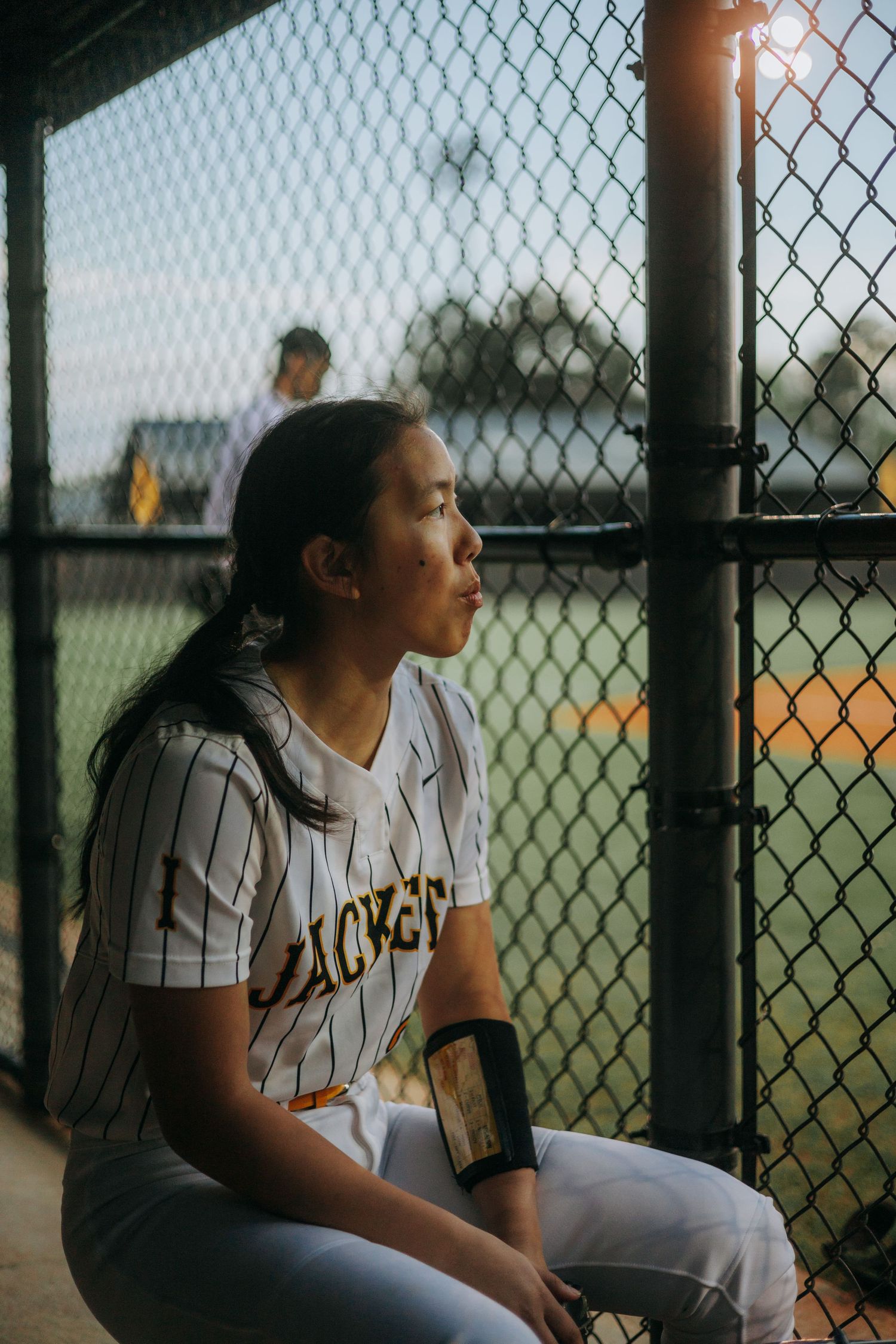 Softball player sitting in dugout looking through chain link fence during game.