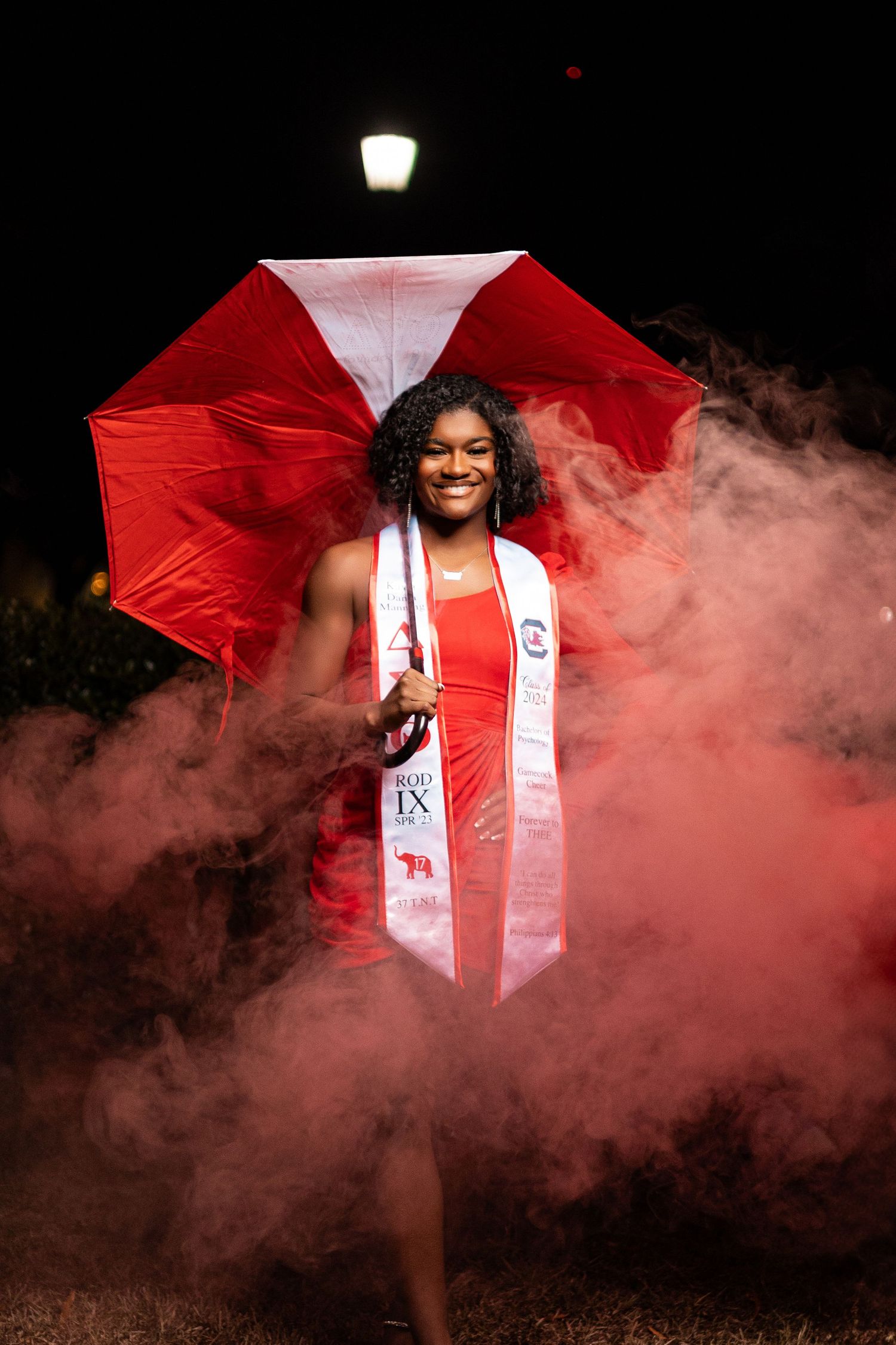 Graduate holding red and white umbrella surrounded by red smoke in dramatic night portrait.