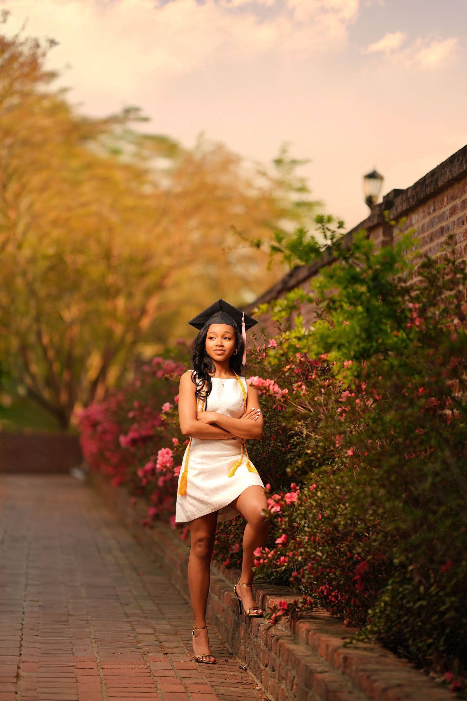 Young woman in white dress and graduation cap poses confidently on brick path surrounded by colorful blooming flowers.