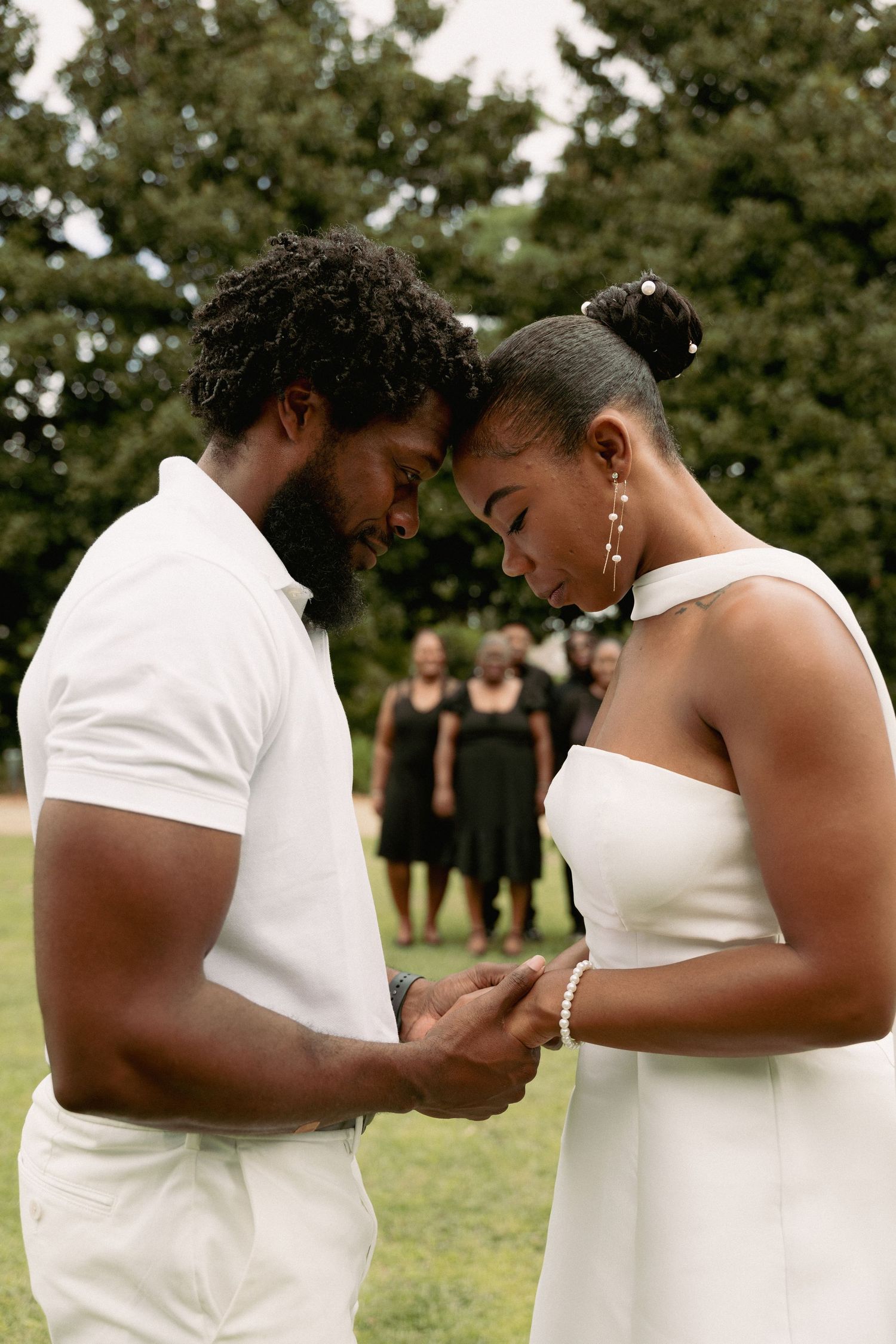 A couple in white attire holds hands and touches foreheads during an outdoor wedding ceremony.