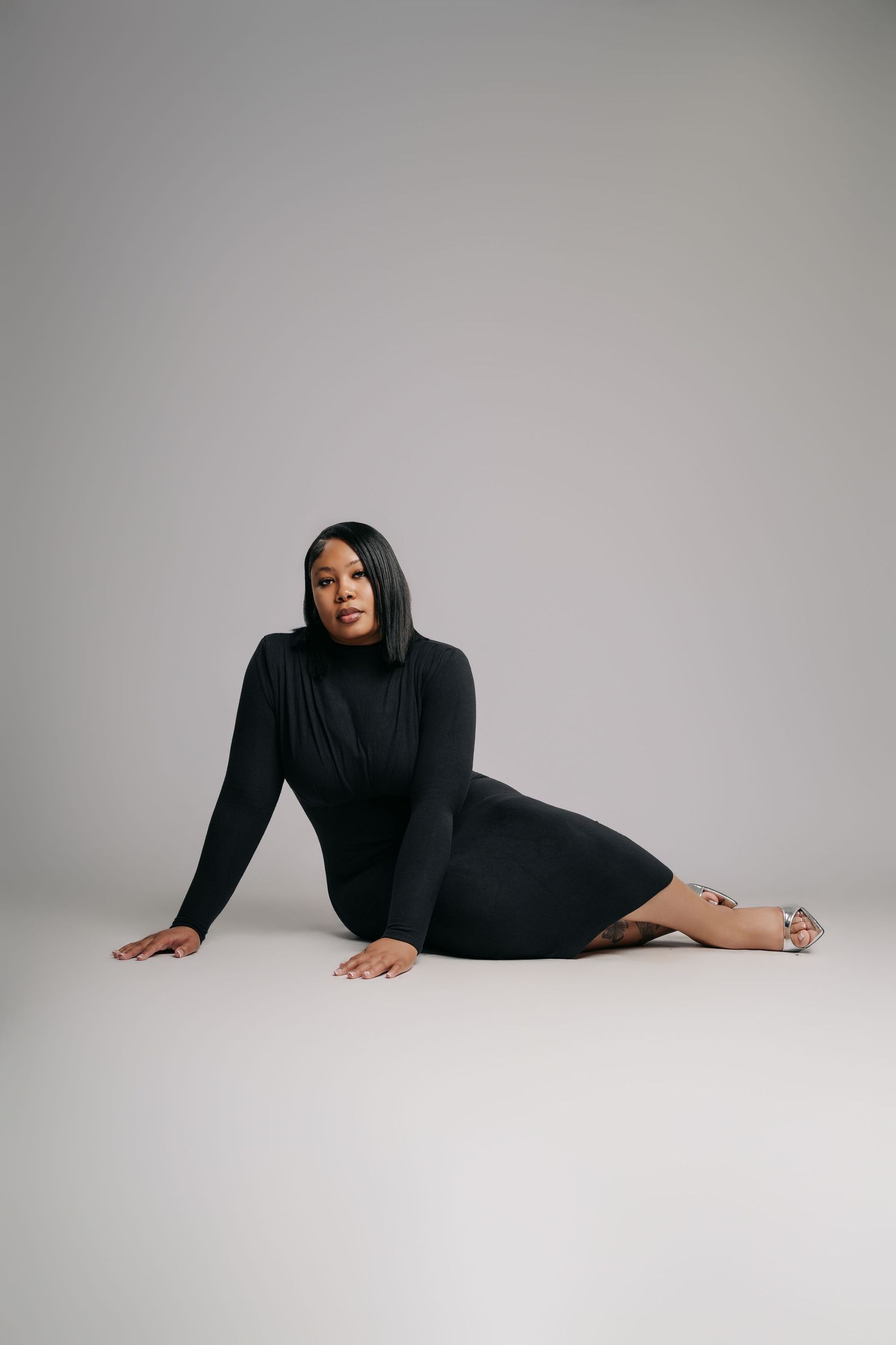 Woman in black turtleneck midi dress posing on floor against gray background.