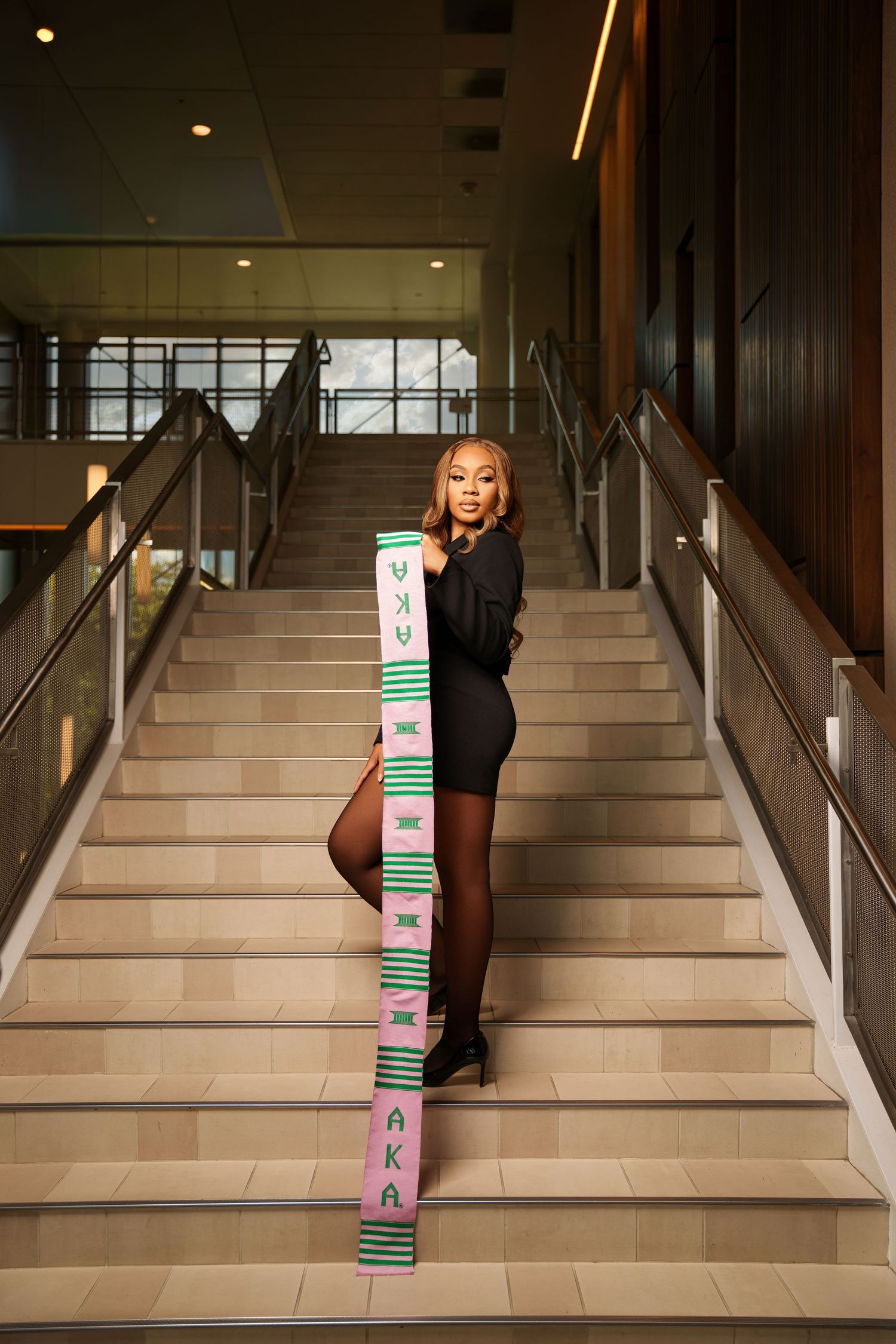 Young woman in black dress poses on stairs holding a pink and green AKA kente cloth stole.