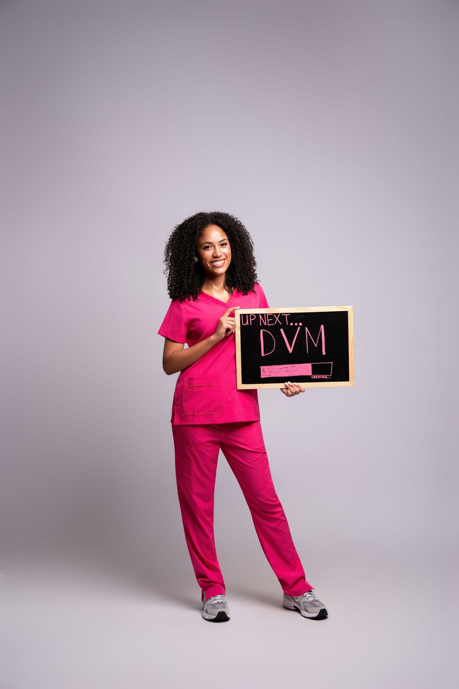 Woman in pink scrubs holding a chalkboard sign reading 'Up Next... DVM' against a gray background.