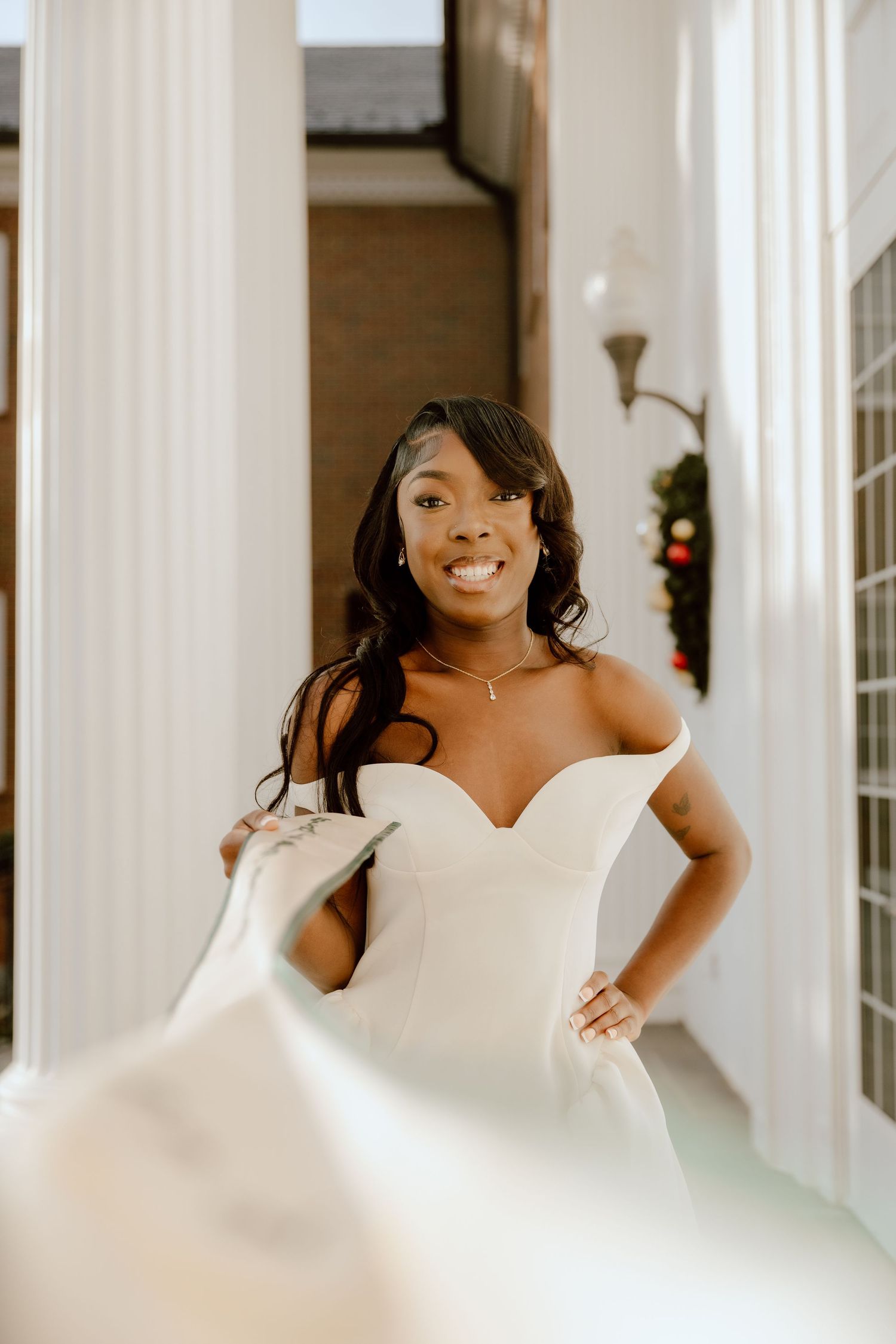 A bride in a white strapless wedding gown poses joyfully in front of white columns and windows.