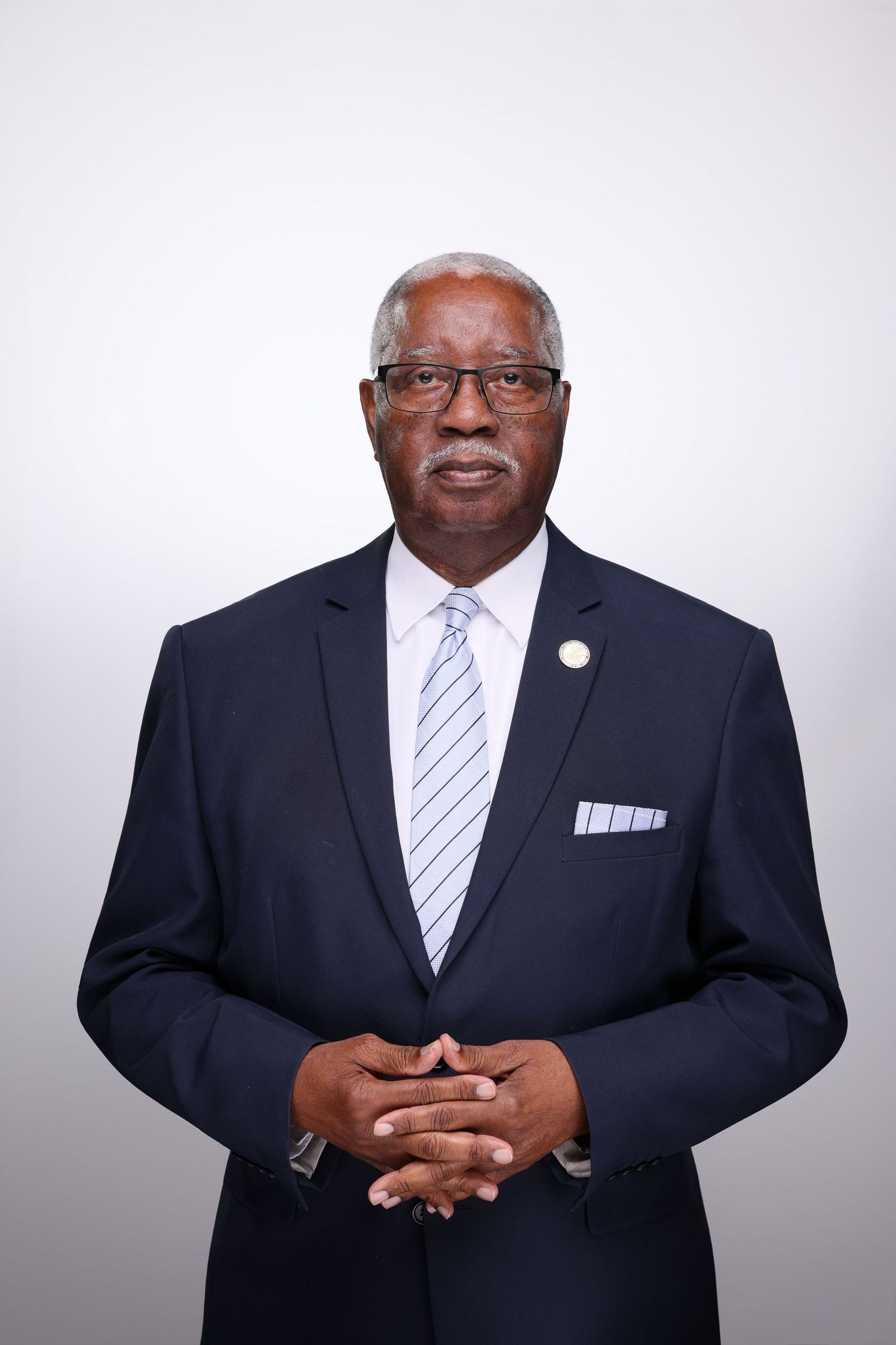 Professional portrait of a senior man in a navy suit with striped tie, glasses, and pocket square, hands clasped.